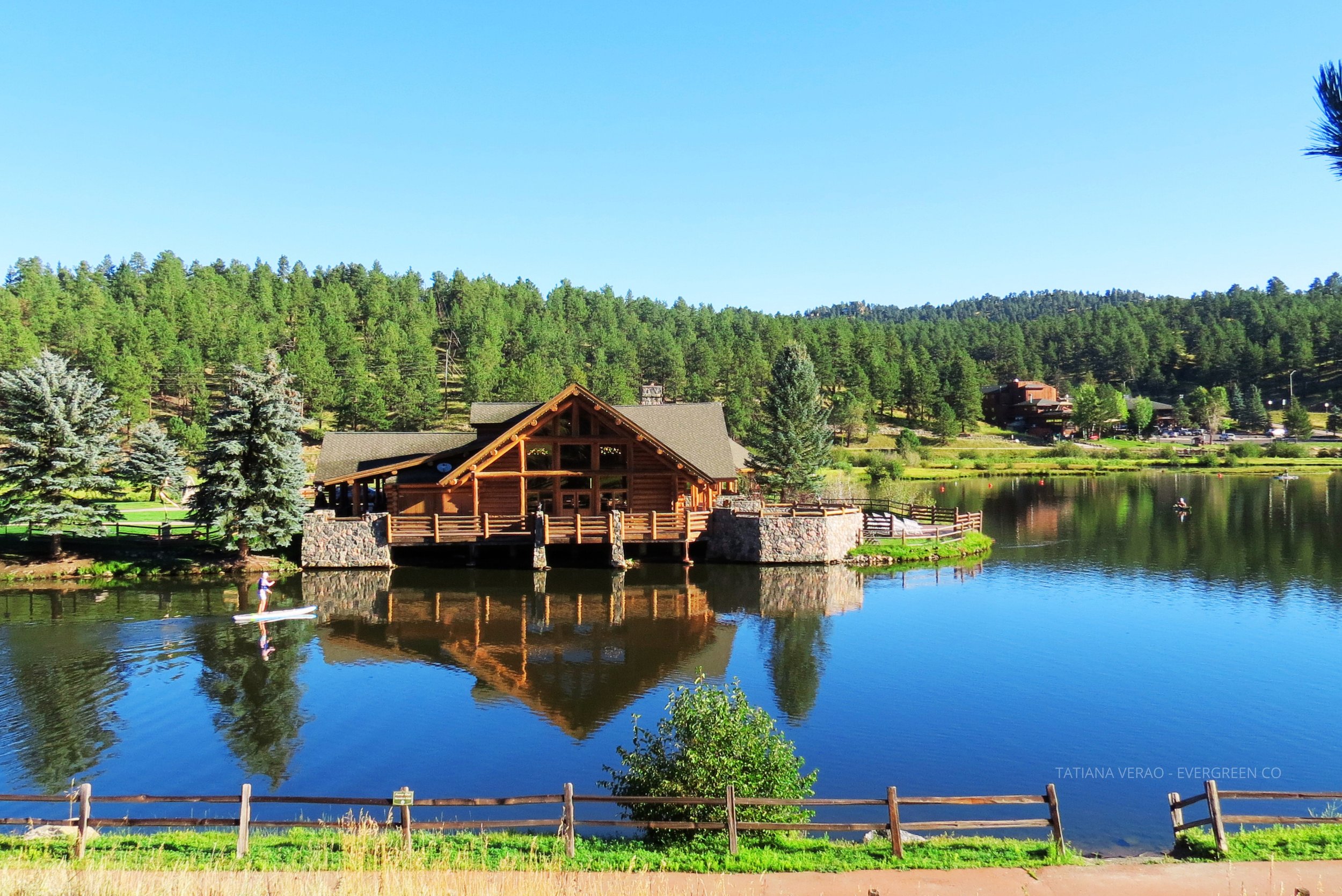A wooden lodge beside a lake with trees and hills in the background, with a person paddleboarding on the water.