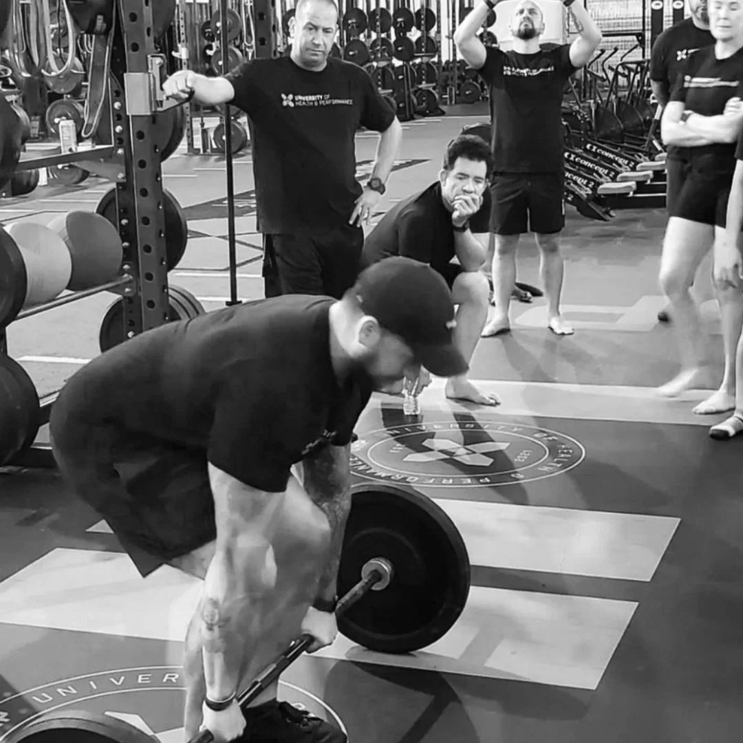 A man in a black t-shirt and shorts is performing a deadlift with a barbell at a gym, surrounded by several people observing, some standing and one squatting with a finger on his chin.