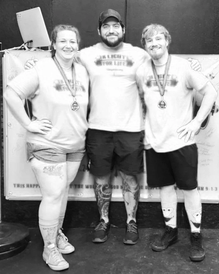 Three people standing together in a gym, wearing medals around their necks. Two of them are women on either side of a man in the middle, all smiling and with their arms around each other. They are dressed in casual athletic clothing and shorts, with tattoos visible on their legs.