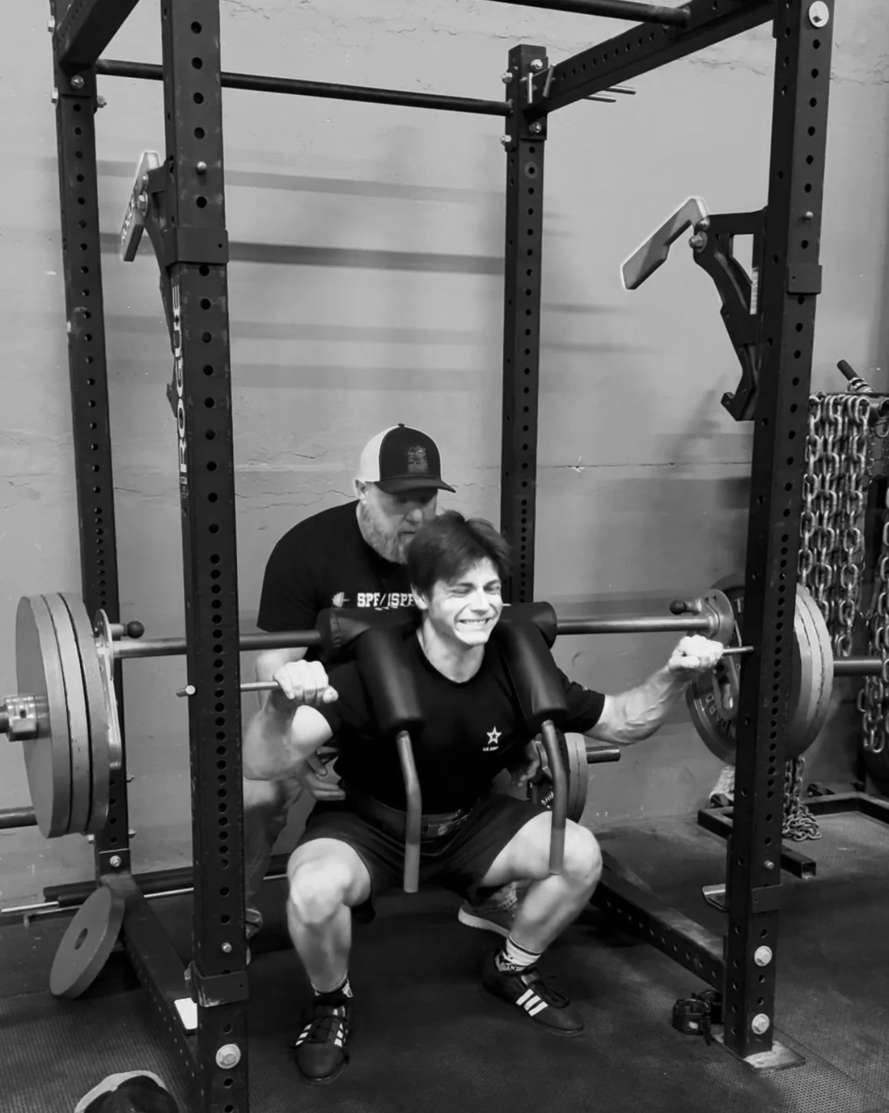 A woman is doing a squat with a barbell loaded with weights, assisted by a man standing behind her at a gym.