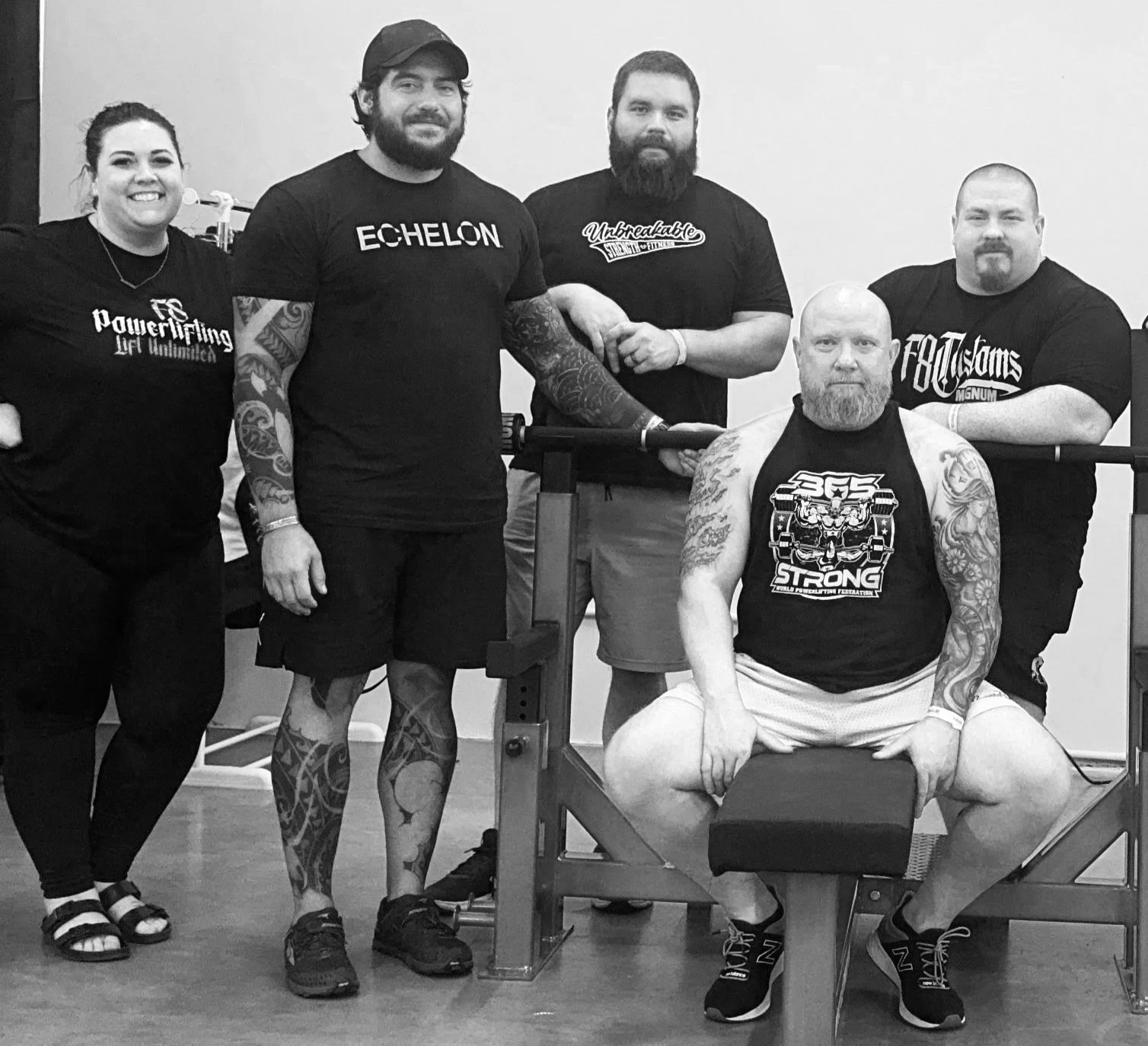 Group of six people posing together in a gym, with some leaning on weights and a bench, all wearing athletic clothing, in a black and white photo.