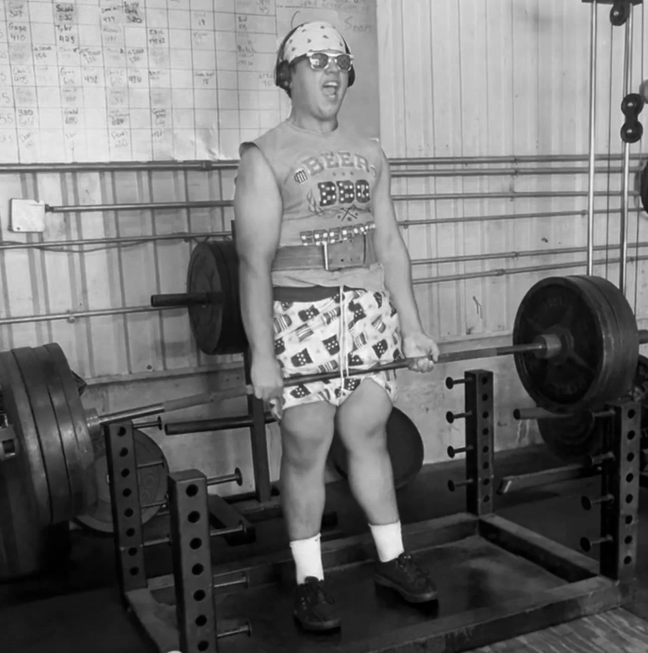 A person lifting a heavy barbell with large weights in a gym. The person is wearing sunglasses, a bandana, a sleeveless shirt, patterned shorts, and socks with shoes.