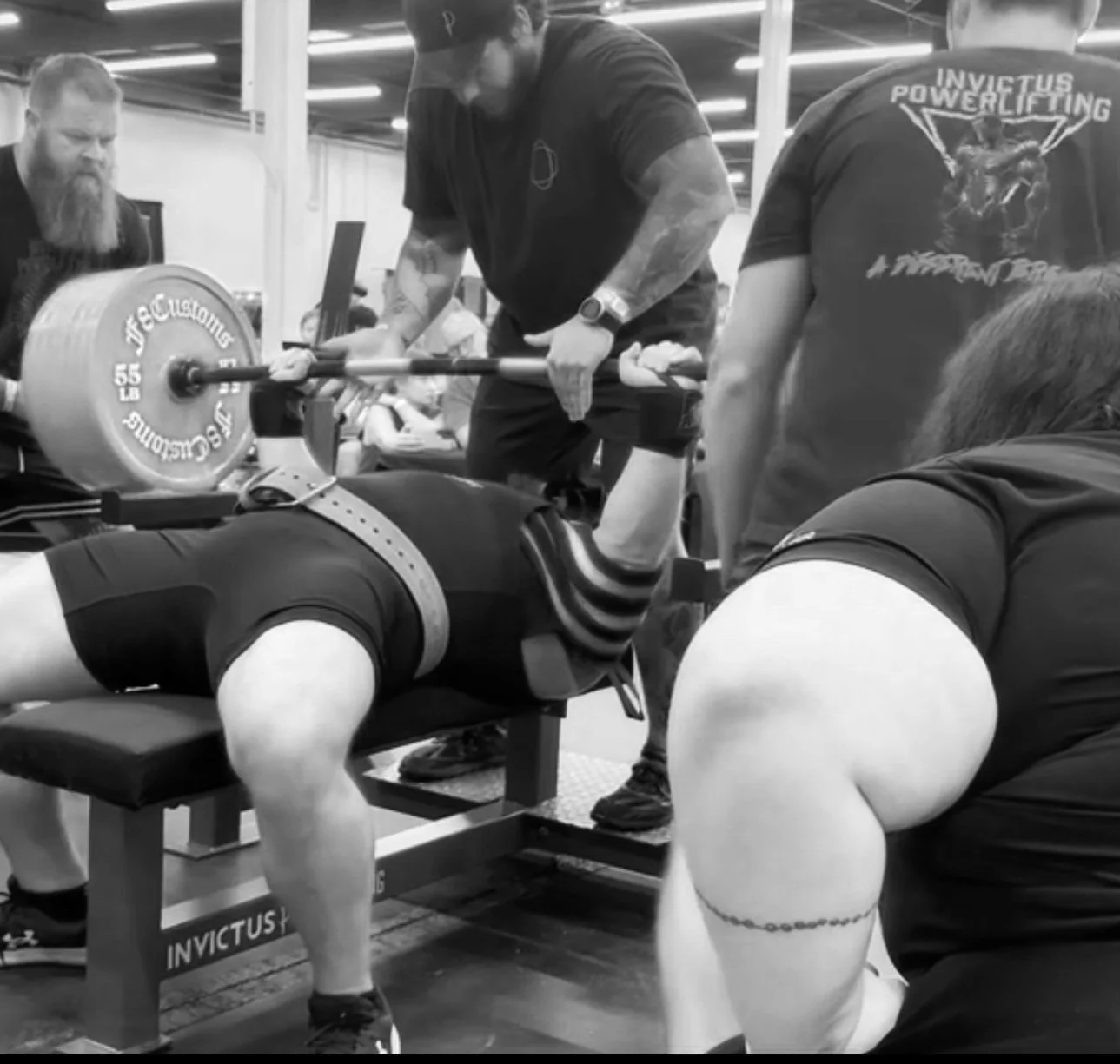 A man performing a bench press at a powerlifting competition, assisted by spotters, with weight plates on the barbell. Others are observing nearby.