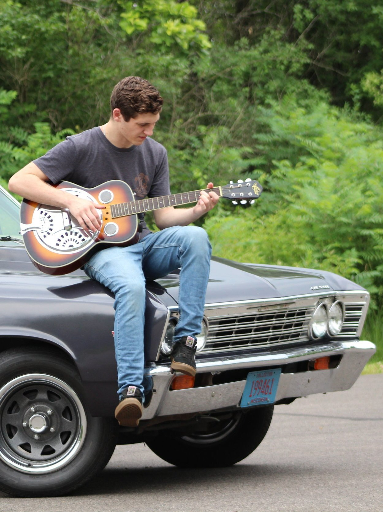 A young man sitting on the front of a black vintage car playing an acoustic guitar, surrounded by green foliage. From Harvard, Illinois in McHenry County doing a senior portrait session. Rush Creek Conservation in Harvard, Illinois.