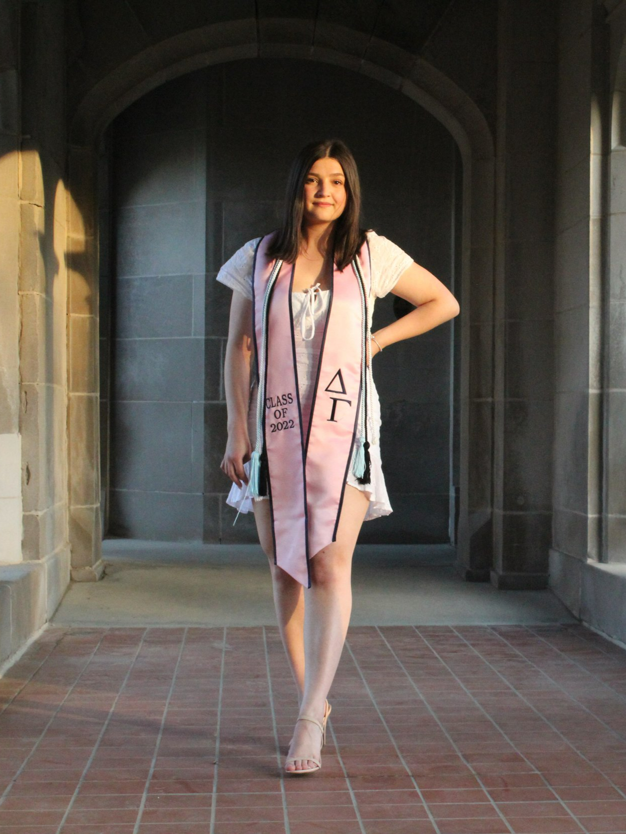 Graduating senior in graduation cap and gown, wearing a pink graduation stole, in a hallway with stone walls and tiled floor. Senior year portrait. Lake Forest College in Lake Forest, Illinois in Lake County. Portrait by Pure Moment Photography.