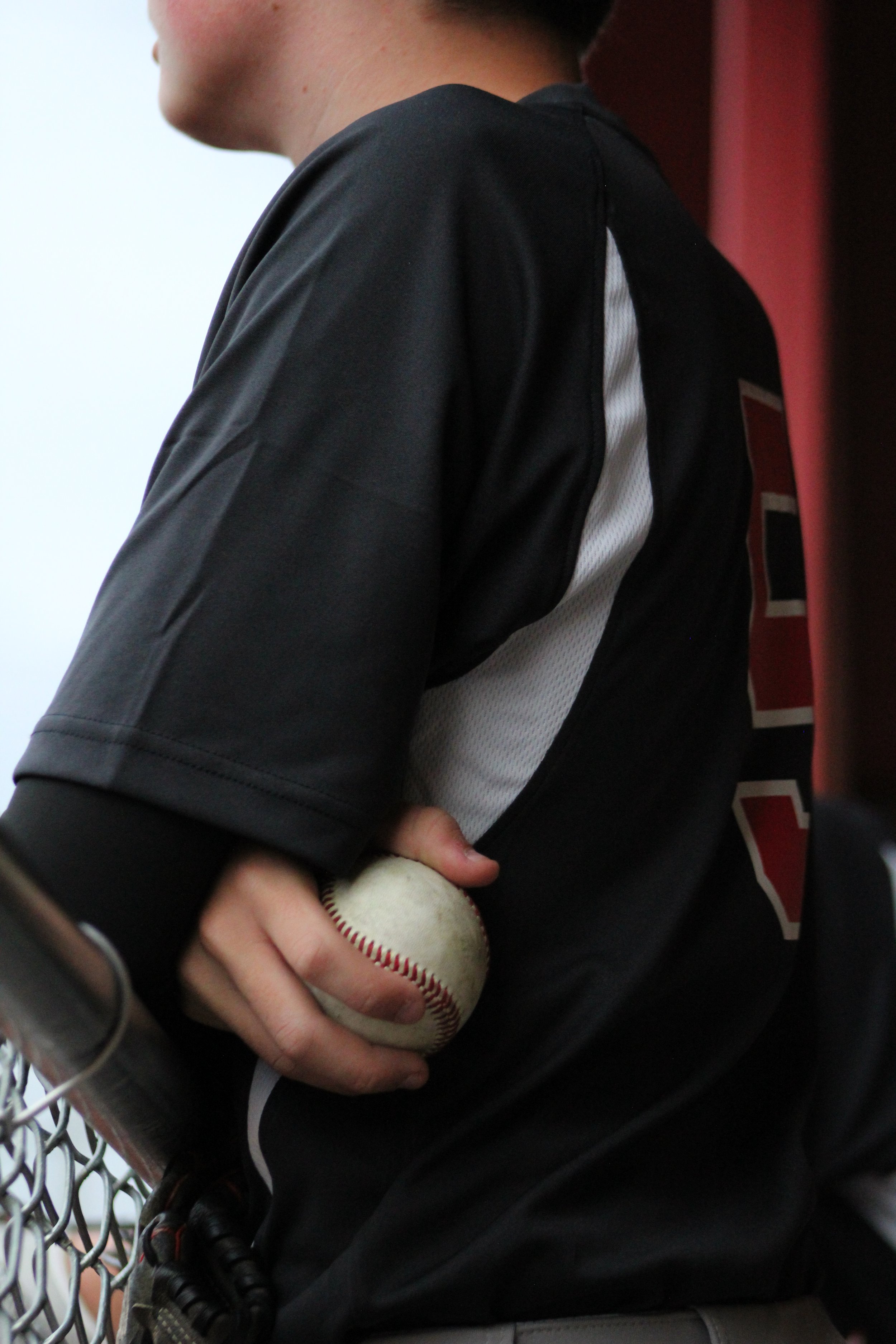 Close-up of a baseball player holding a ball while leaning on a dugout fence in Walworth, Wisconsin captured during a local game at Big Foot High School. Natural, candid sports photography by Pure Moment Photography.