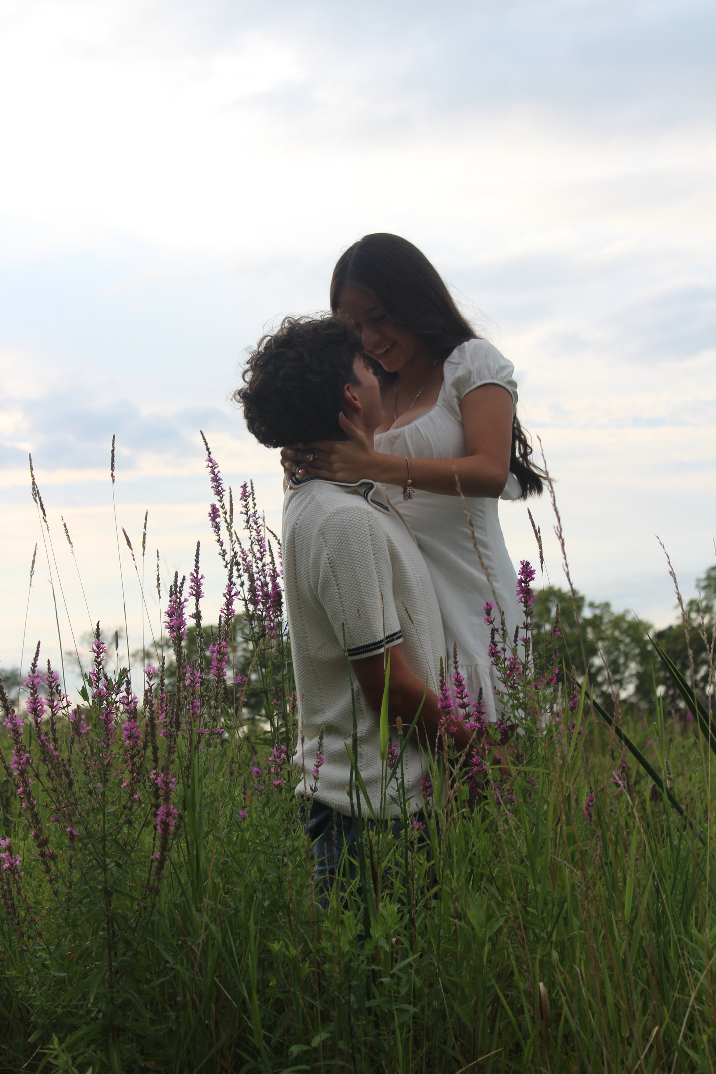 A couple standing in a field of tall purple flowers, embracing and smiling at each other during sunset or dusk. Couple photoshoot in McHenry, Illinois in McHenry County. Stickney Run Conservation Area, McHenry, Illinois.