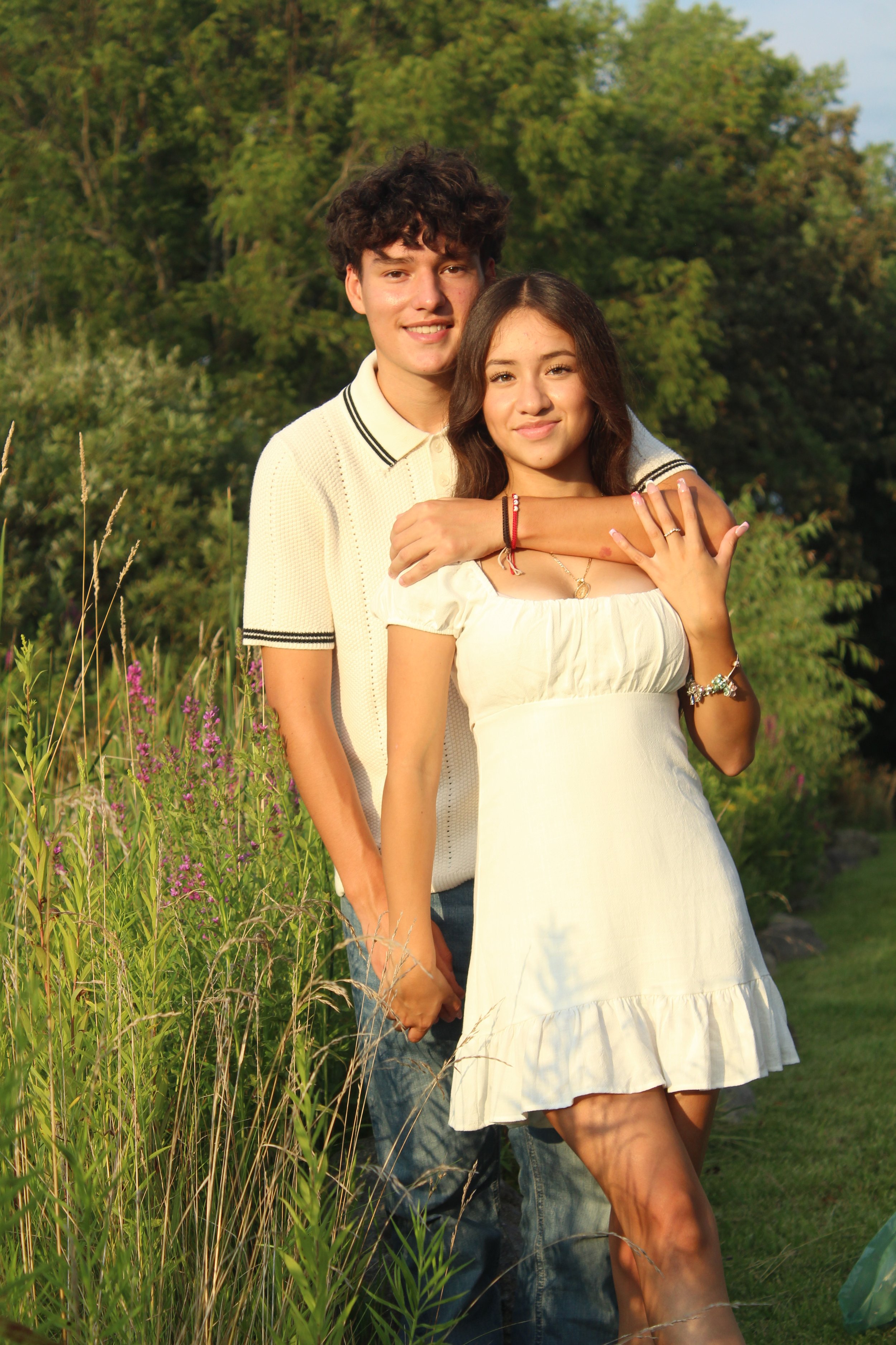A young couple standing outdoors in a field of tall grass and purple flowers, with trees in the background, smiling at the camera. High school graduates taking senior year photos. McHenry, Illinois in McHenry County. Photos by Pure Moment Photography