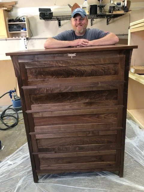 Man with a beard and a cap leaning on a tall wooden piece of furniture in a workshop.