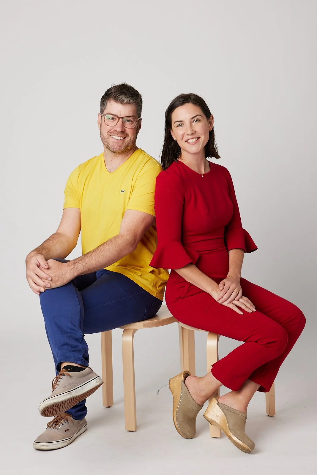 A man and woman sit on wooden stools against a plain white background. The man is wearing a yellow T-shirt, blue pants, and beige shoes. The woman is wearing a red dress and beige high-heeled shoes. They are smiling and sitting with their backs touching.