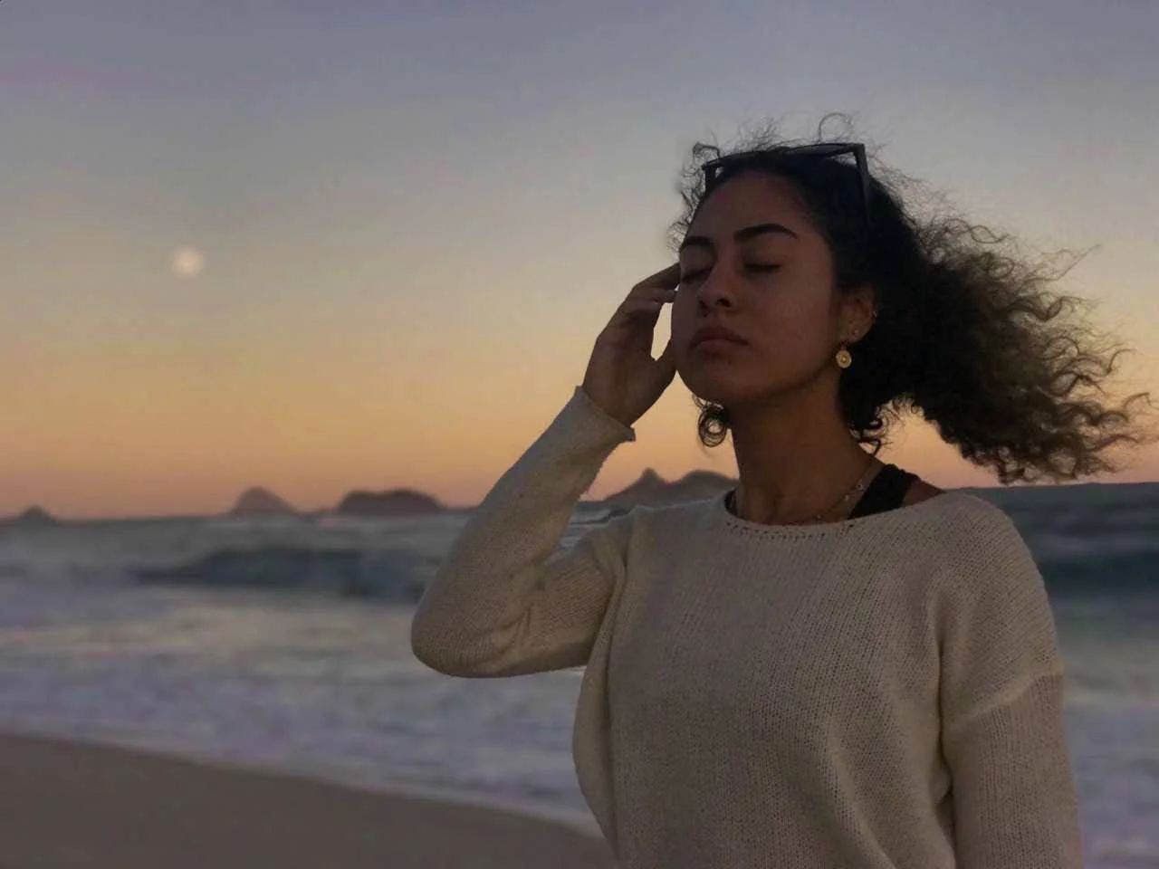 A woman with curly hair standing on the beach during sunset with her eyes closed, one hand touching her face, wearing a beige sweater and sunglasses on her head.