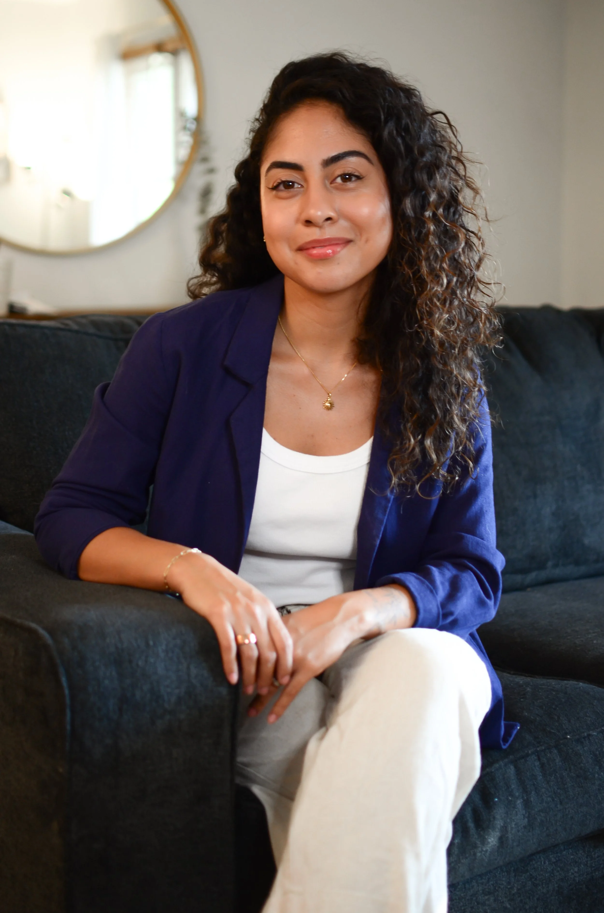 A woman with curly dark hair, wearing a navy blazer and white top, sitting on a black sofa in a cozy, well-lit room.