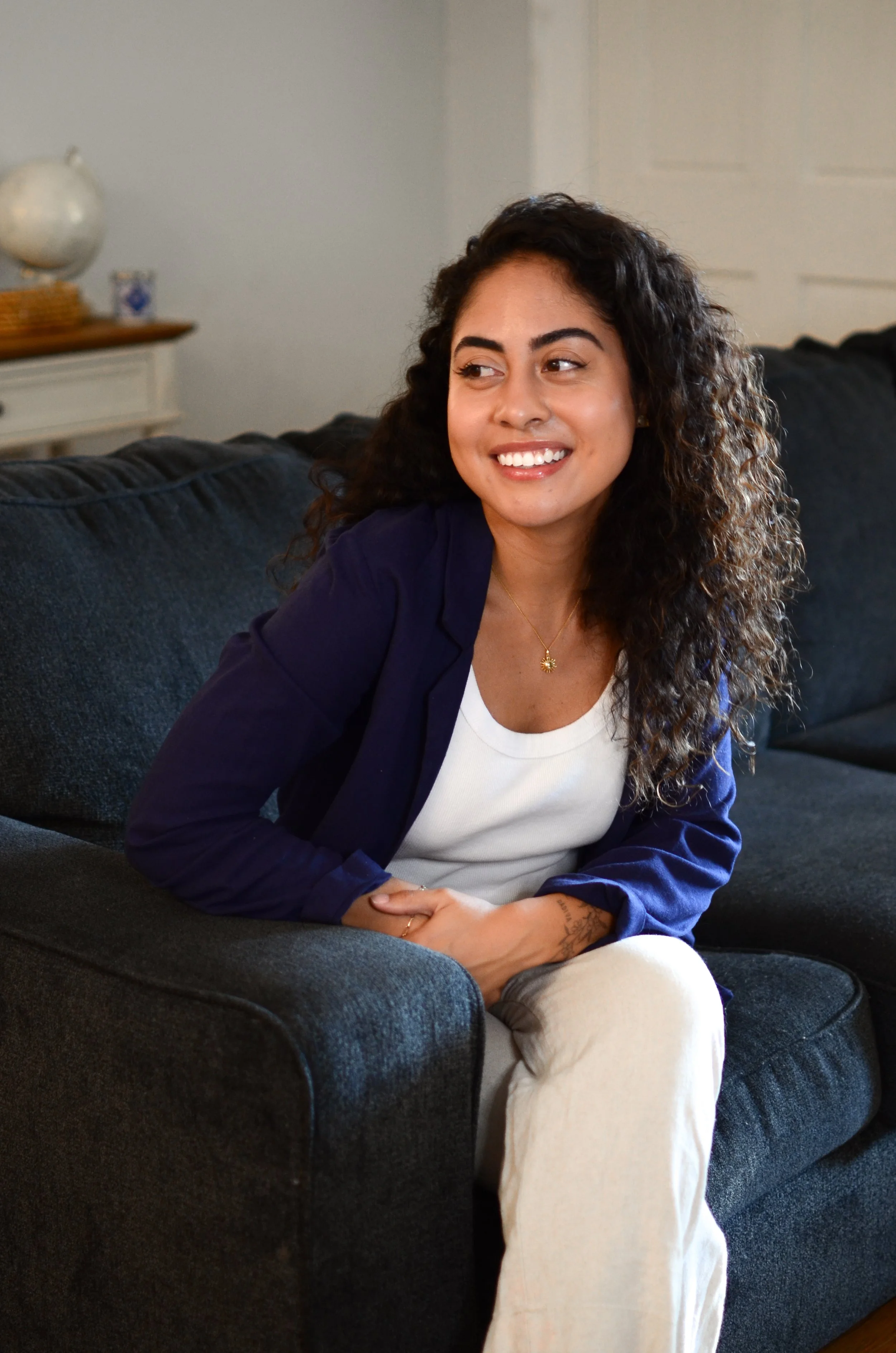 A woman with curly dark hair sitting on a dark sofa, smiling and looking to her left, wearing a white shirt and navy blazer.