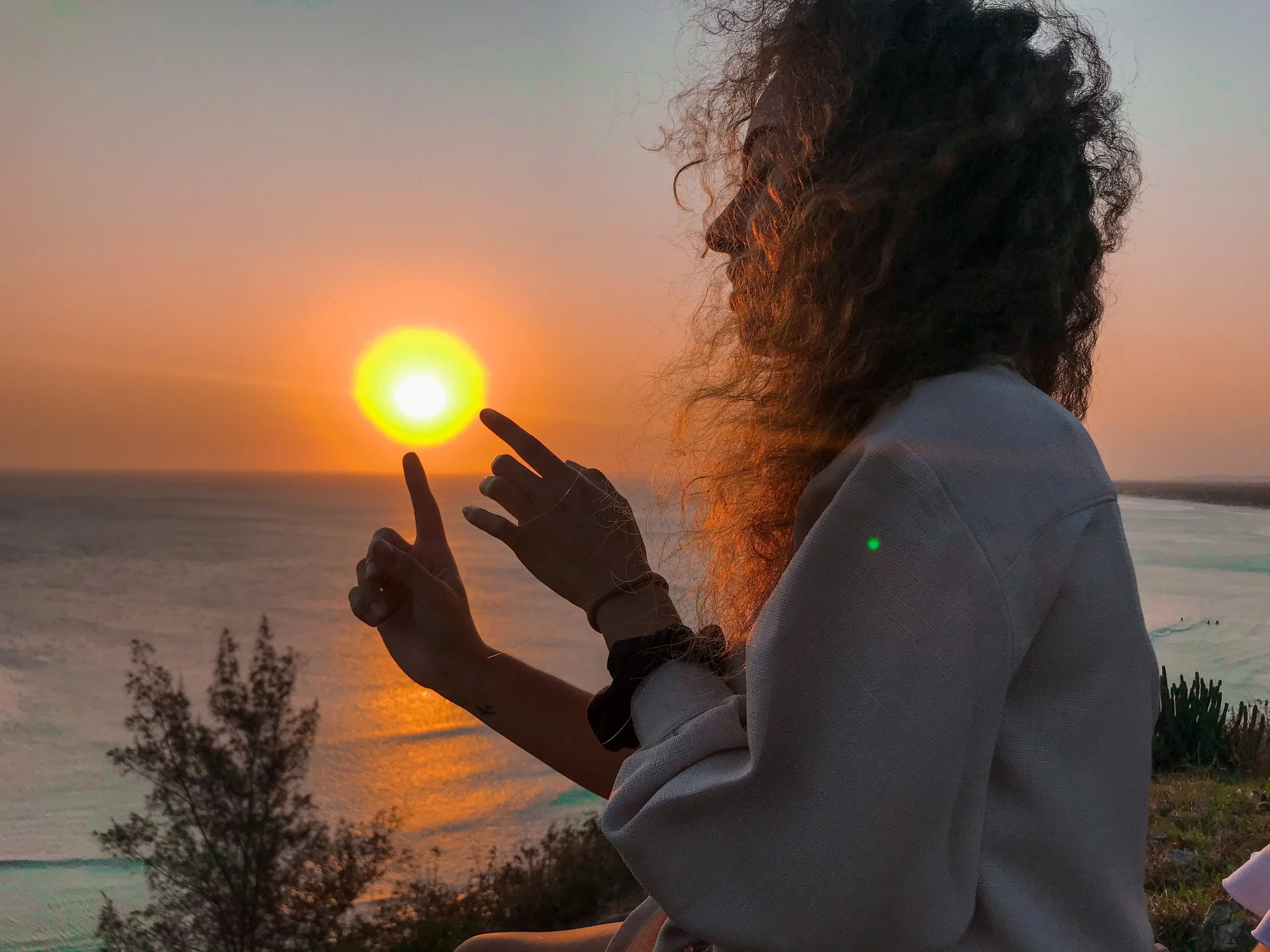 A woman with curly hair touching the setting sun at the beach during sunset.