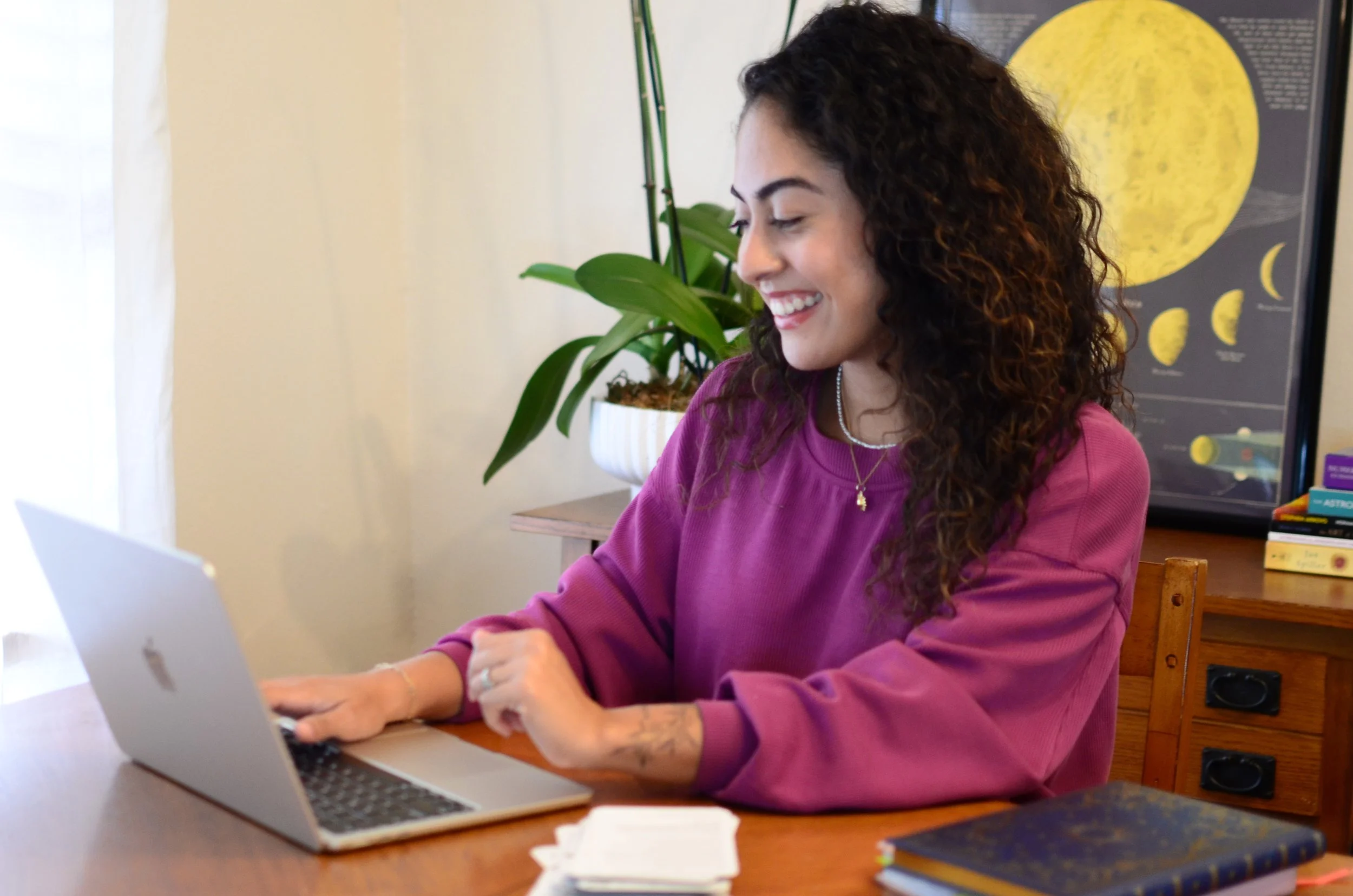 A woman with curly hair wearing a pink sweatshirt and a necklace, smiling while working on a silver MacBook at a wooden desk, with a green potted plant and a lunar poster in the background.