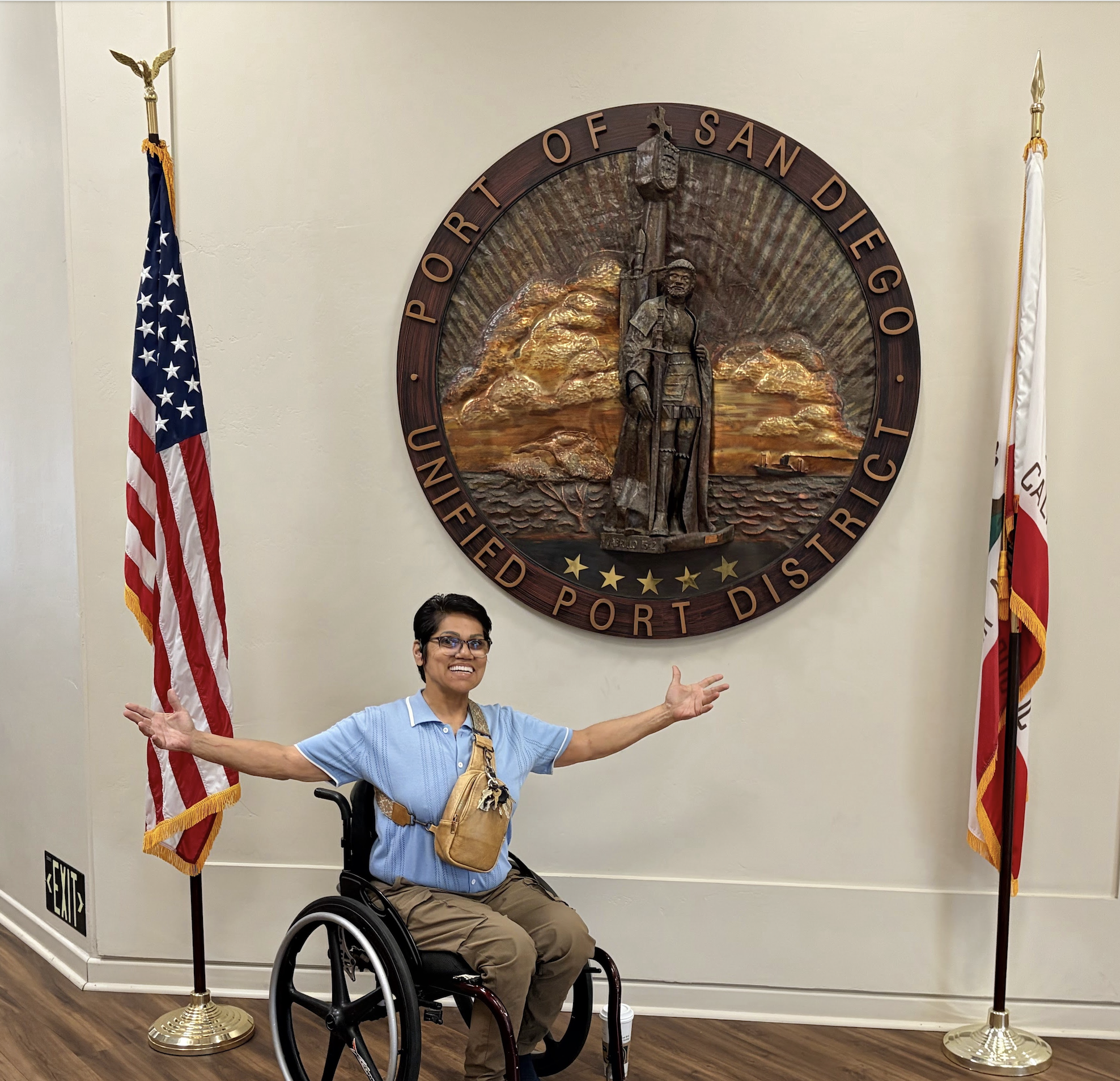 A person in a manual wheelchair poses cheerfully with open arms in front of the official round wooden seal of the Port of San Diego. They are positioned inside the Port Administration Building, bordered by two standing flags.