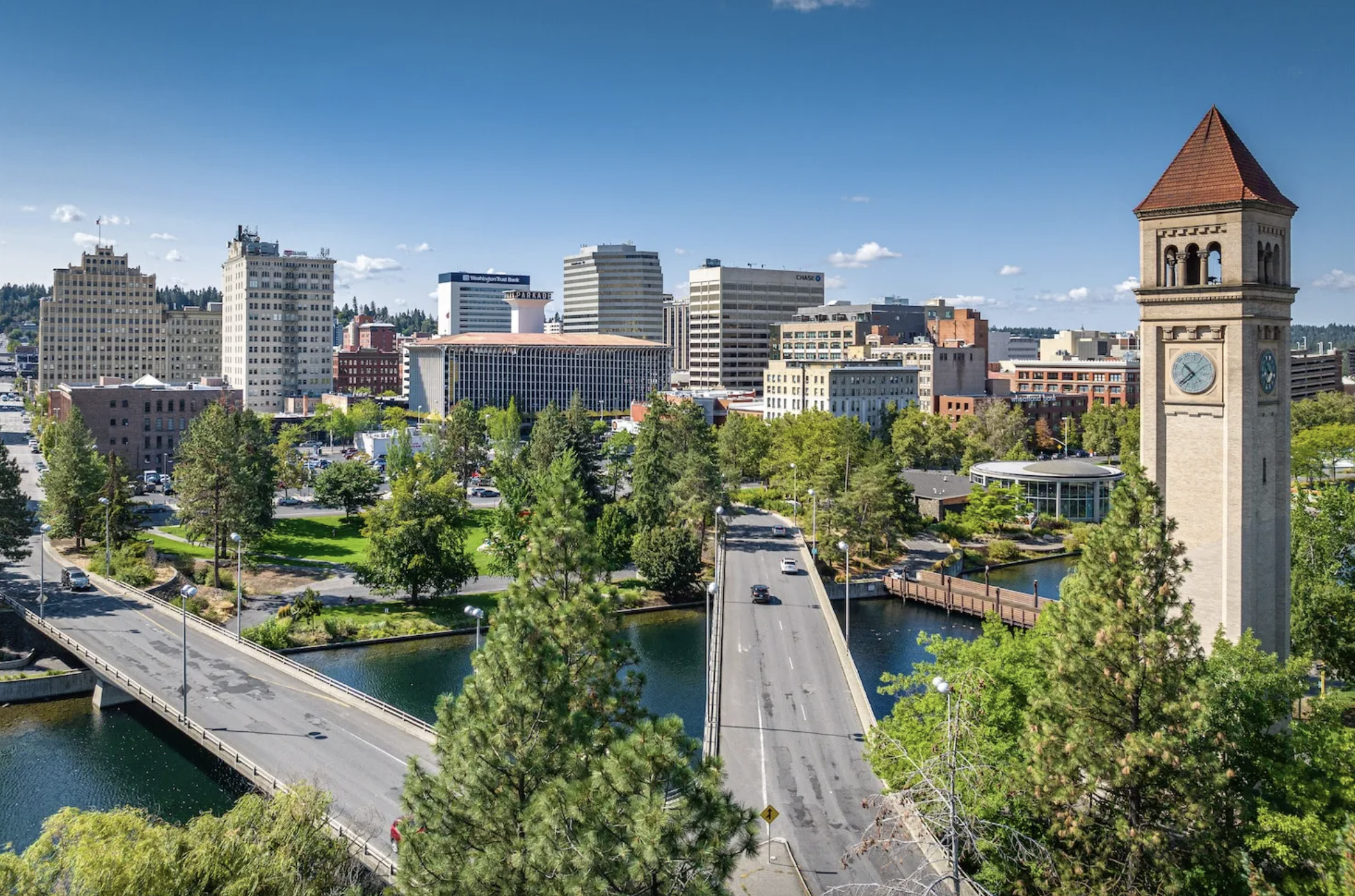 City skyline with a clock tower, a river, and a bridge, surrounded by trees and urban buildings under a blue sky.