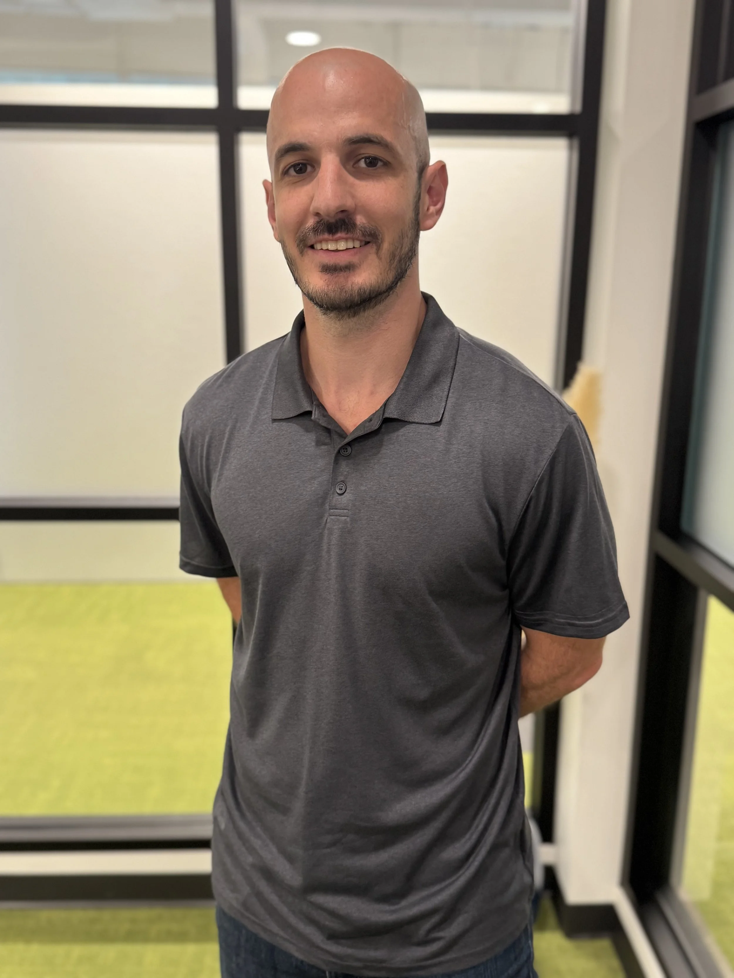 A young man with a shaved head and beard, wearing a gray polo shirt, standing indoors near a glass wall with a yellow-green carpet in the background.