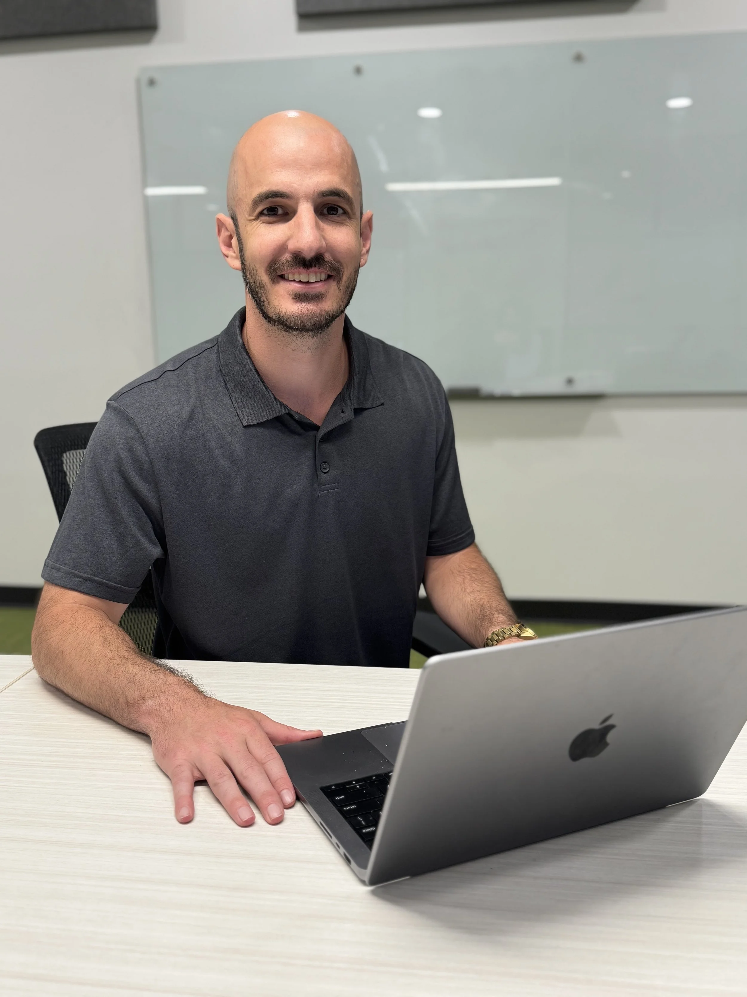 A man sitting at a desk with a silver MacBook laptop, smiling. He has a bald head, a beard, and is wearing a dark gray polo shirt. Behind him is a whiteboard in a classroom or office setting.