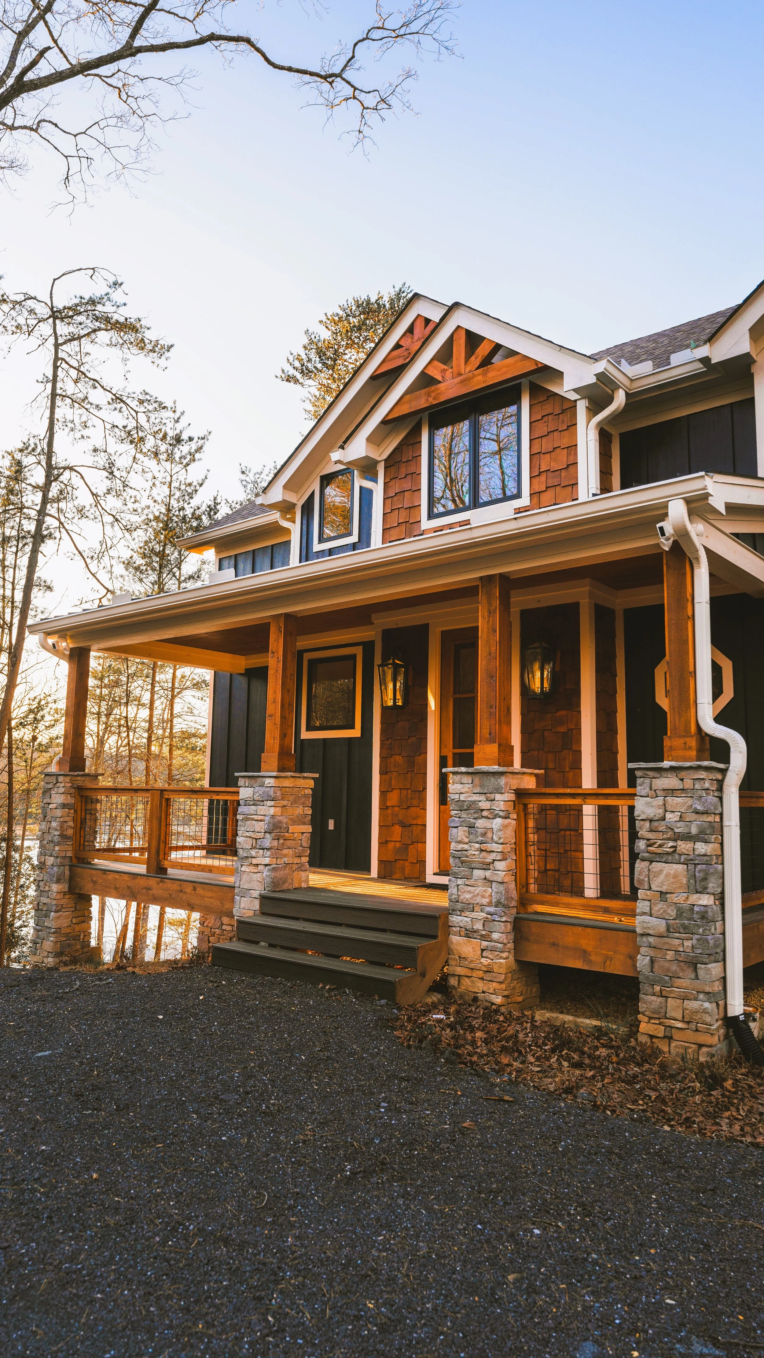 Front view of a custom modern farmhouse two-story house georgia with a porch, wooden and stone columns, on a slightly elevated black gravel yard with trees in the background.