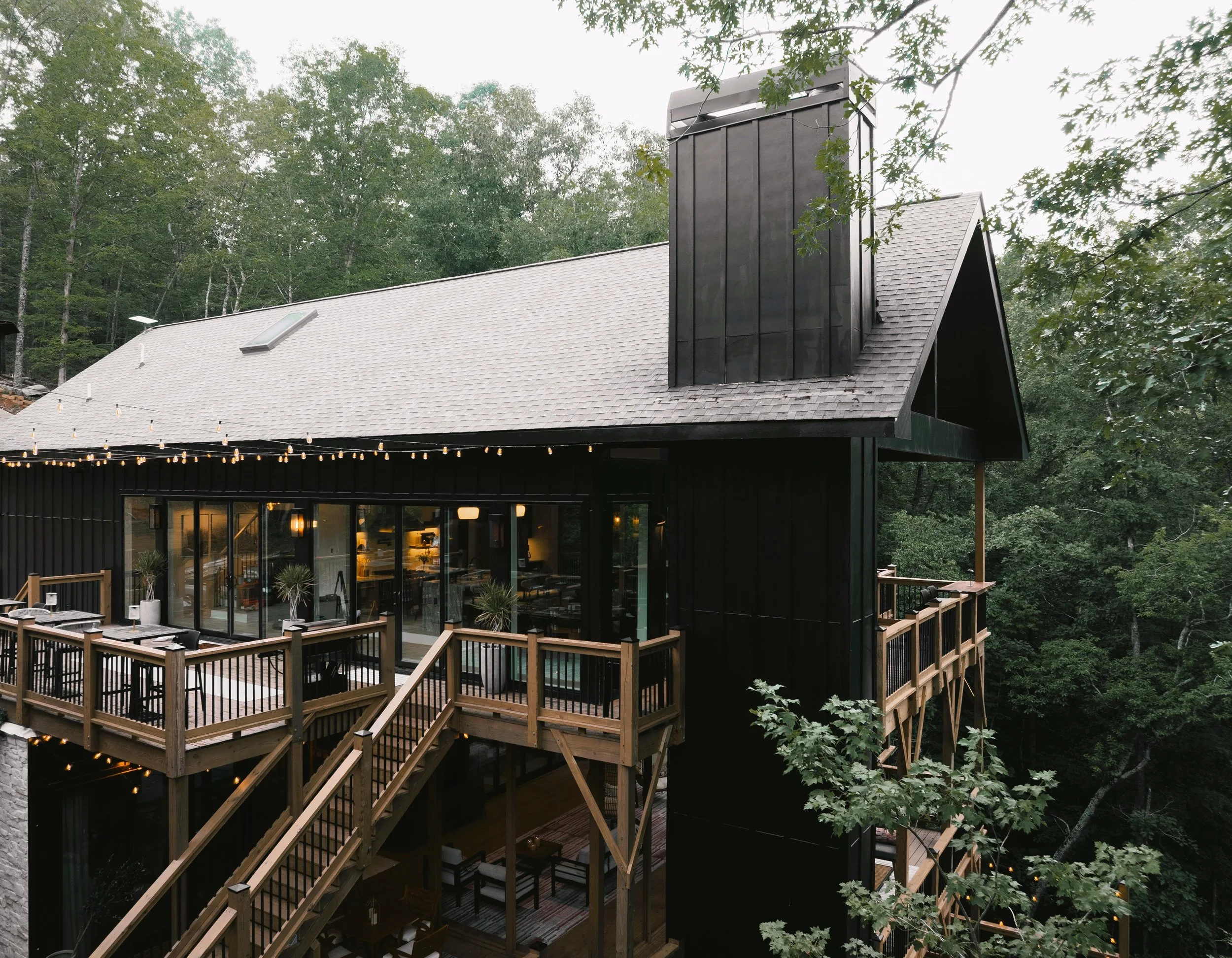 A modern black house with a wooden deck, large glass doors, and string lights, set in a forested area.