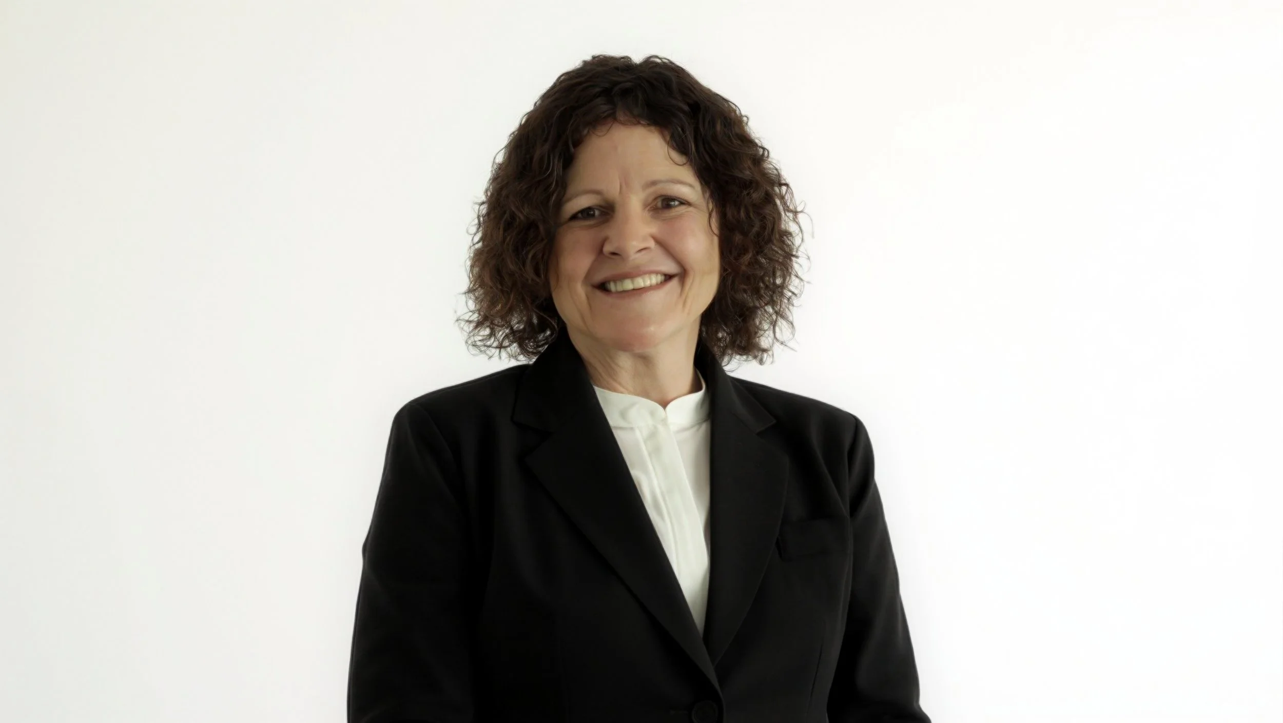 A woman with curly brown hair wearing a black blazer and white blouse, smiling against a plain white background.