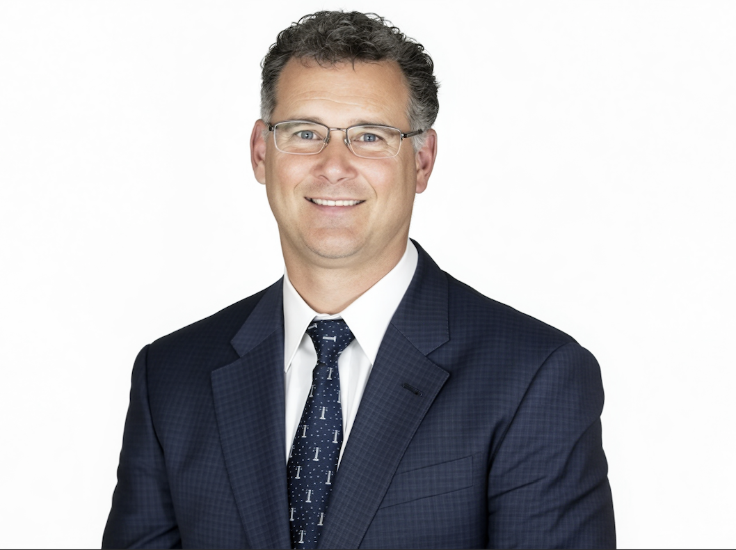 Professional headshot of a middle-aged man with glasses, wearing a navy blazer, white dress shirt, and a navy tie, smiling against a plain white background.