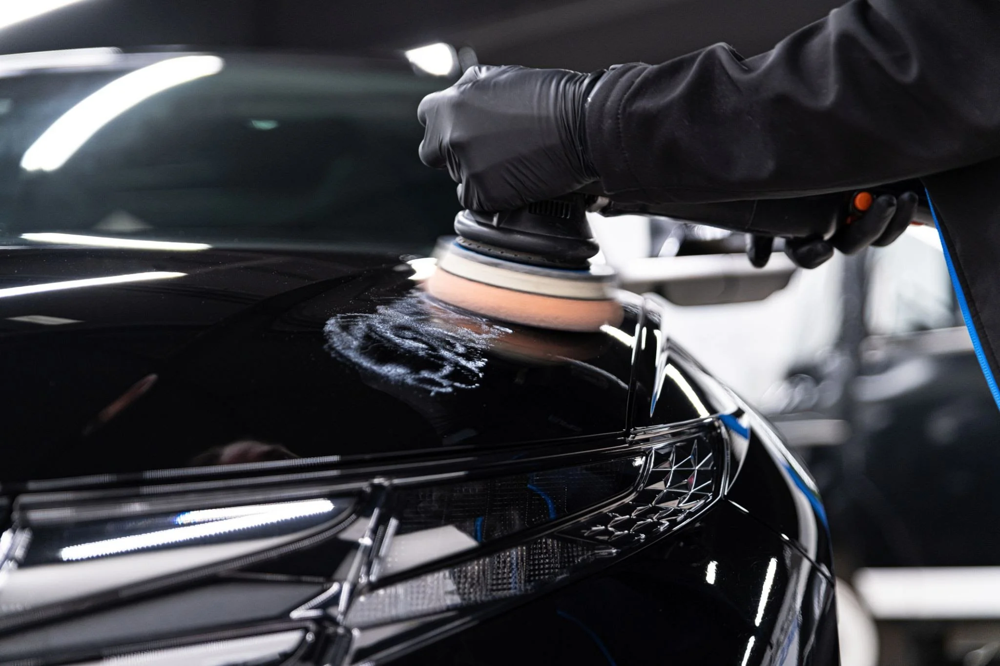 A person wearing black gloves and a black jacket is polishing a black car with a polishing machine inside a garage or workshop.
