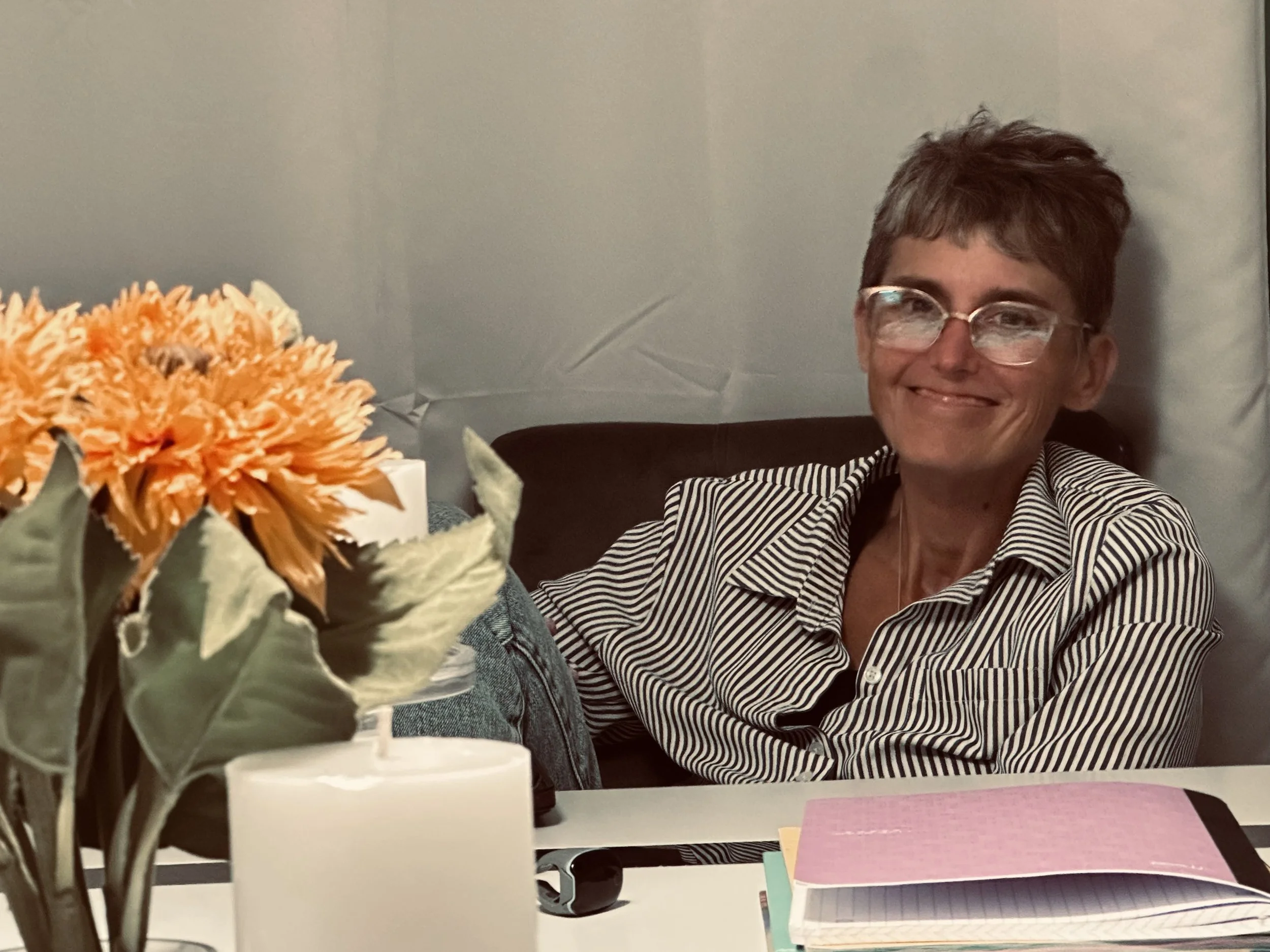 A woman with short brown hair and glasses, smiling, sitting at a table. In the foreground, there is a bouquet of orange flowers and some colorful notepads on the table. The background has a plain light gray wall.