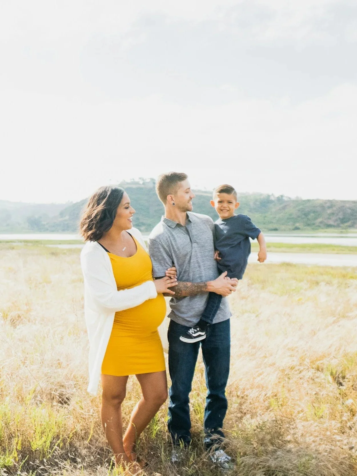 Family of three: pregnant woman in yellow dress, man holding young boy, standing in golden field near water with hills in background.