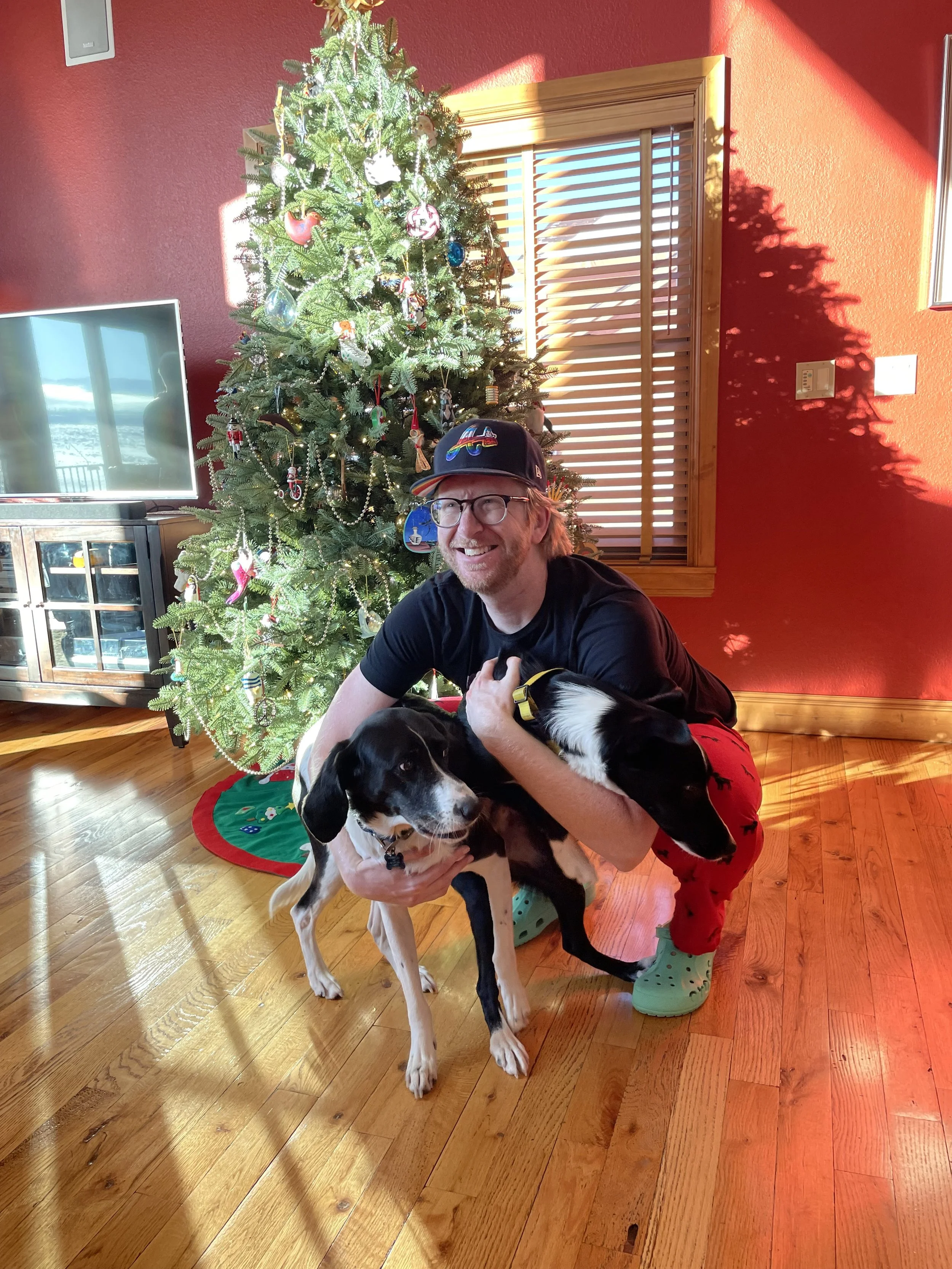 Eric smiling and kneeling on the wooden floor while holding two black and white dogs in front of a decorated Christmas tree, with a red wall and a window behind him.