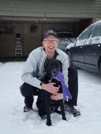 Eric, wearing a gray jacket and black pants, kneeling on snow outside a garage, smiling and hugging a black dog with a purple and blue bandana.