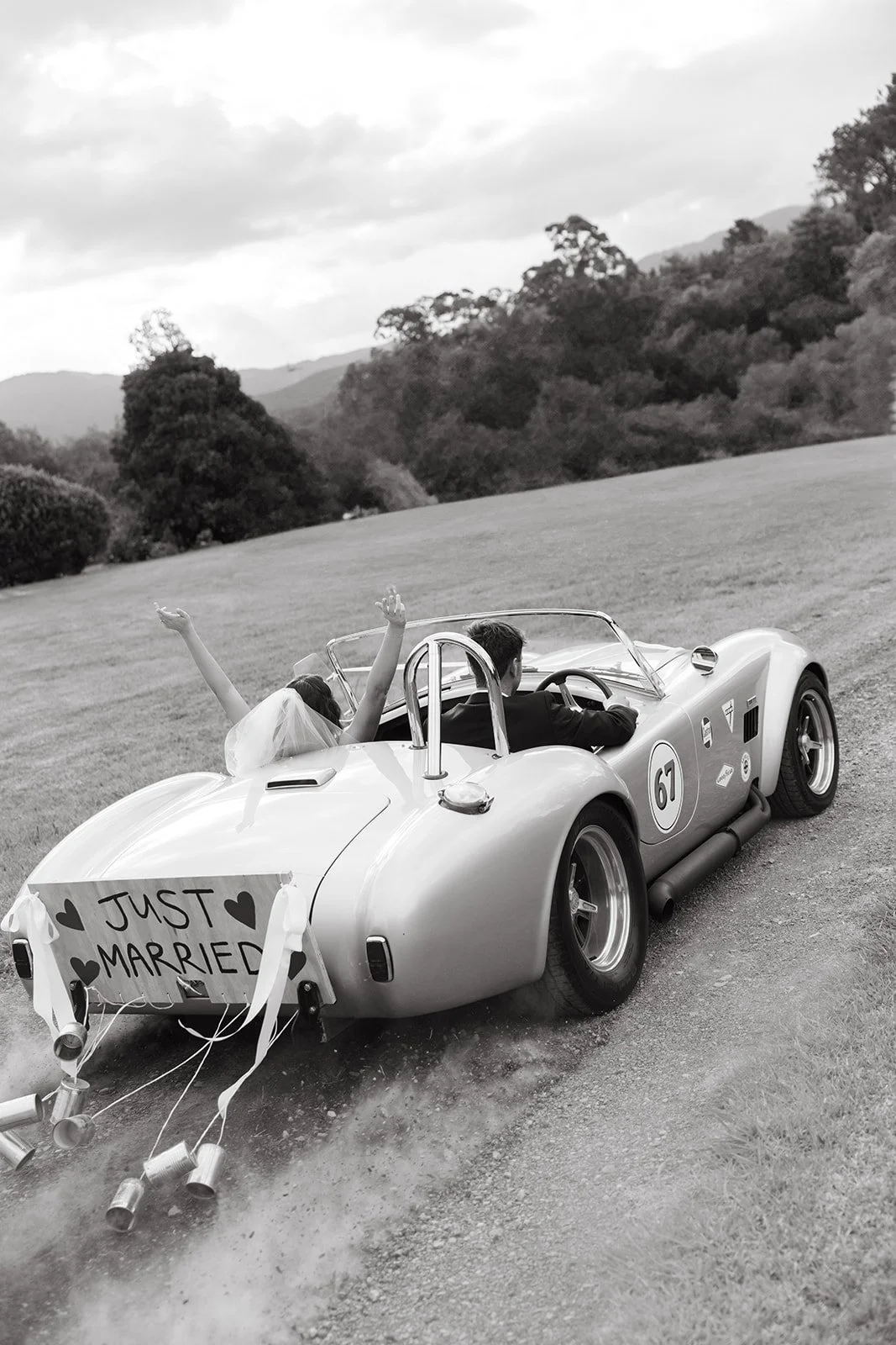 A newlywed couple in a vintage race car with a 'Just Married' sign on the back, driving on a dirt road with trees and hills in the background.