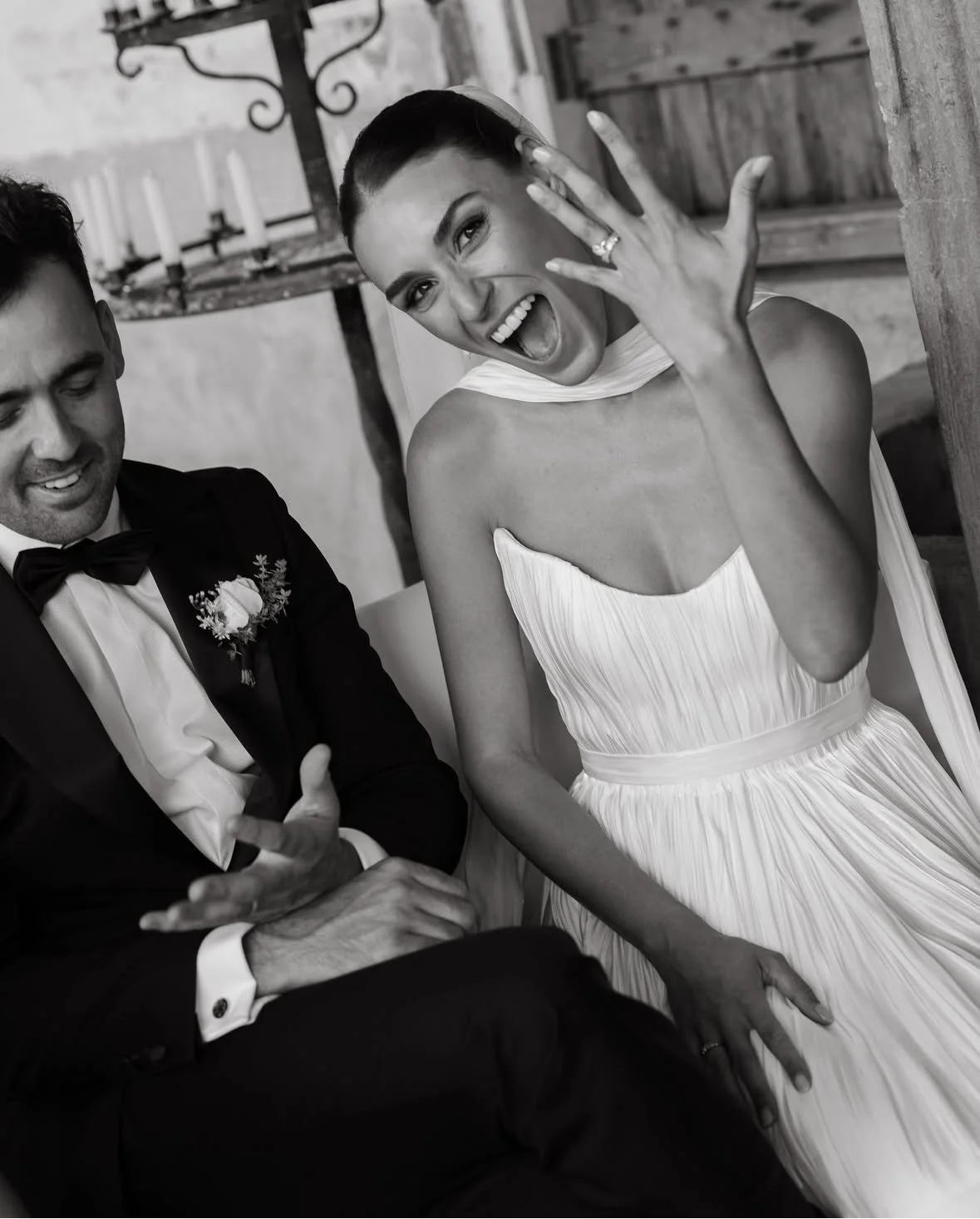 A bride in a white dress showing a joyful expression and hand gesture, sitting beside a groom dressed in a tuxedo with a flower boutonniere, during a wedding celebration.