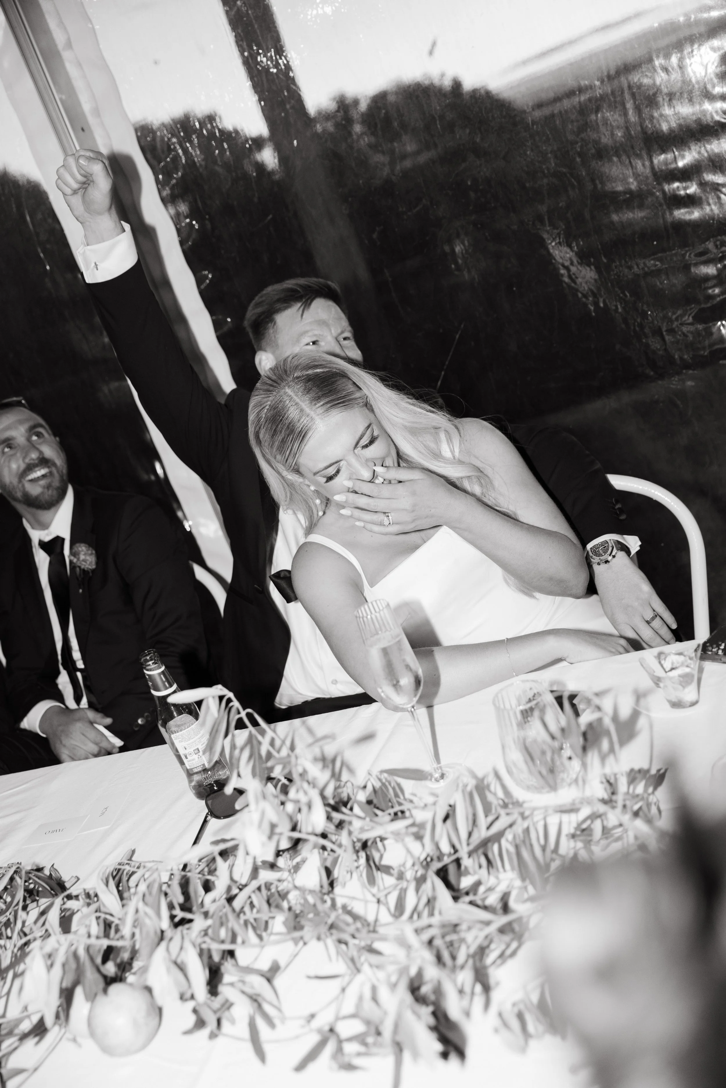 A wedding reception scene in black and white with a bride and groom laughing, the groom raising a fist, and other guests sitting at a decorated table with flowers, champagne, and drinks.