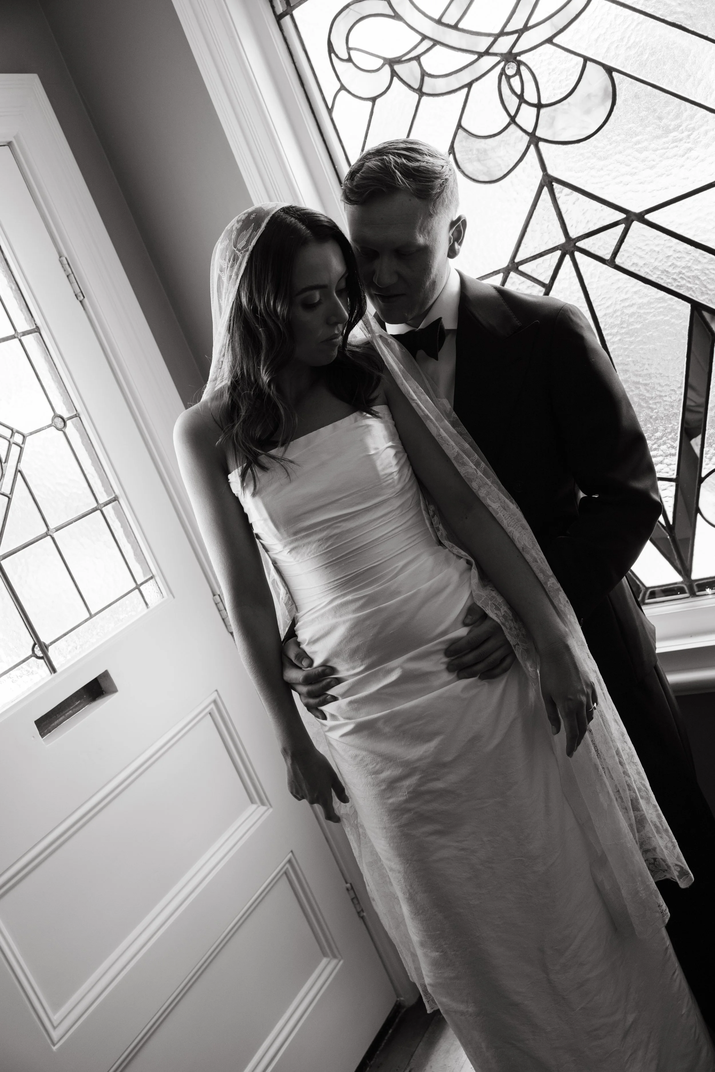 A bride and groom standing close together indoors near a stained glass window, with the bride wearing a strapless wedding dress and veil, and the groom in a tuxedo. The photo is in black and white.