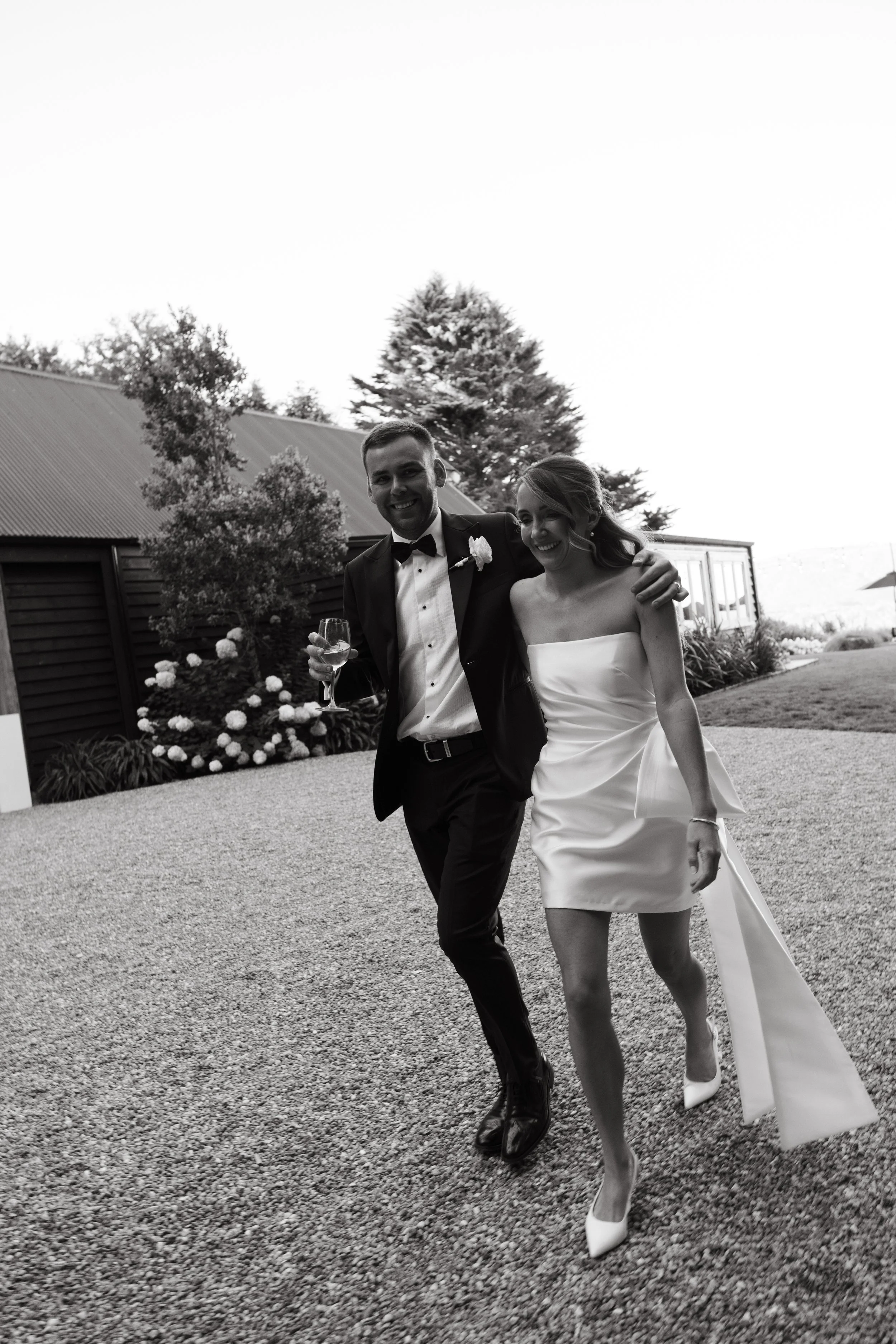 Black and white photo of a happy couple in wedding attire, walking outdoors, with the man holding a glass of wine and smiling, and the woman in a strapless wedding dress, smiling and walking with her arm around him.