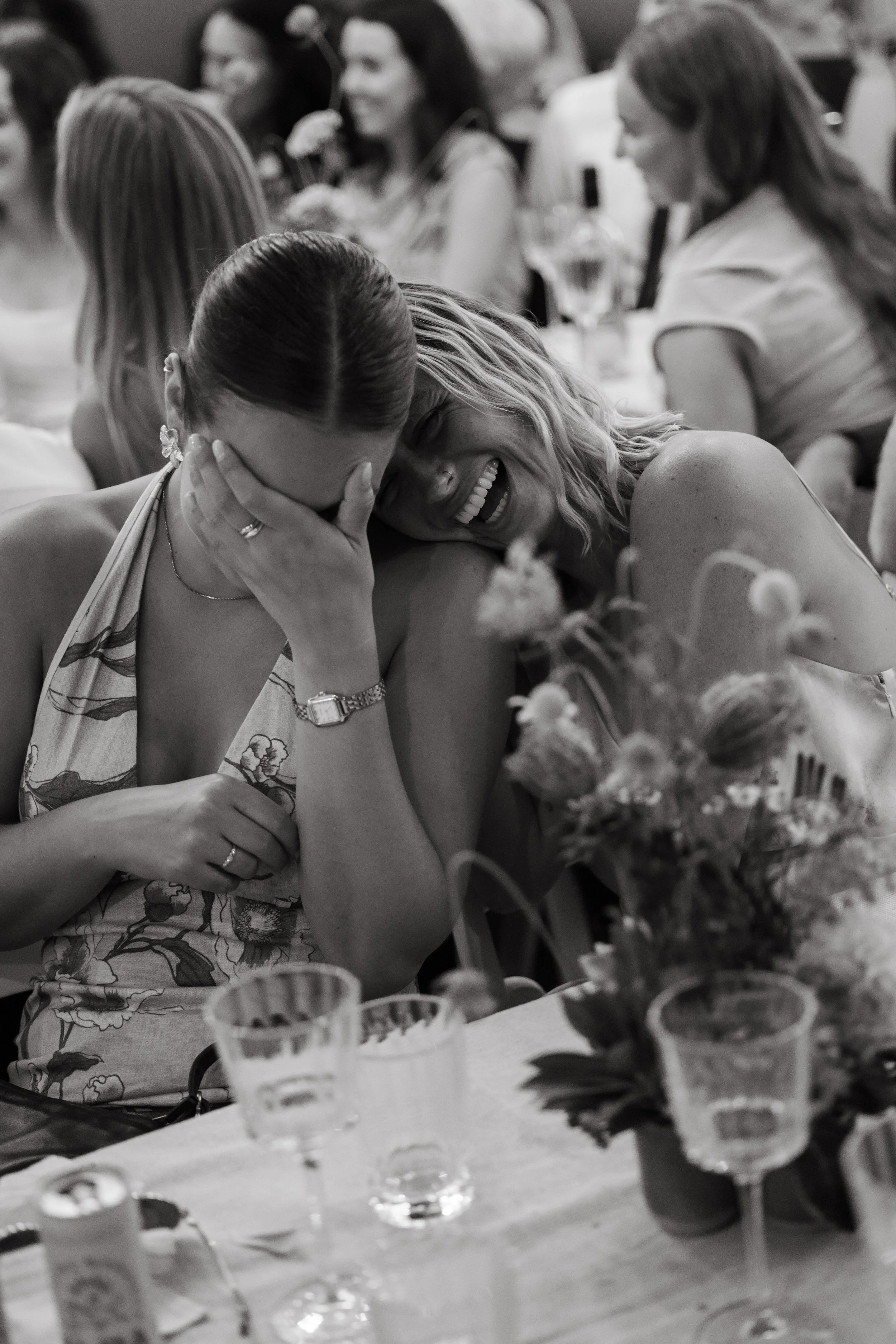 Two women sharing a laugh at a social gathering, with one covering her face and the other smiling broadly.