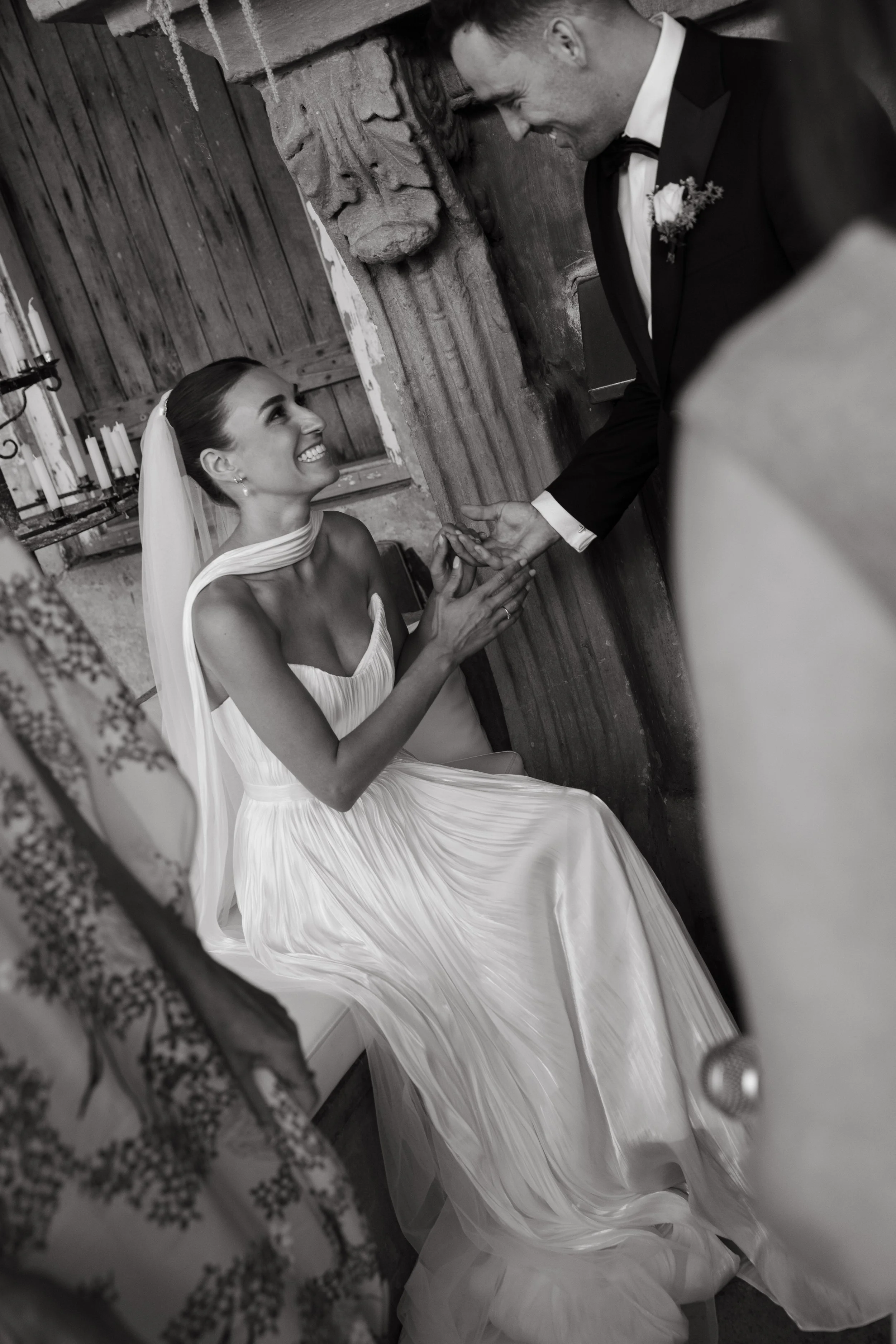 A bride and groom during their wedding ceremony, with the bride seated and smiling at the groom who is holding her hand, in a rustic setting with exposed wood wall and candles.
