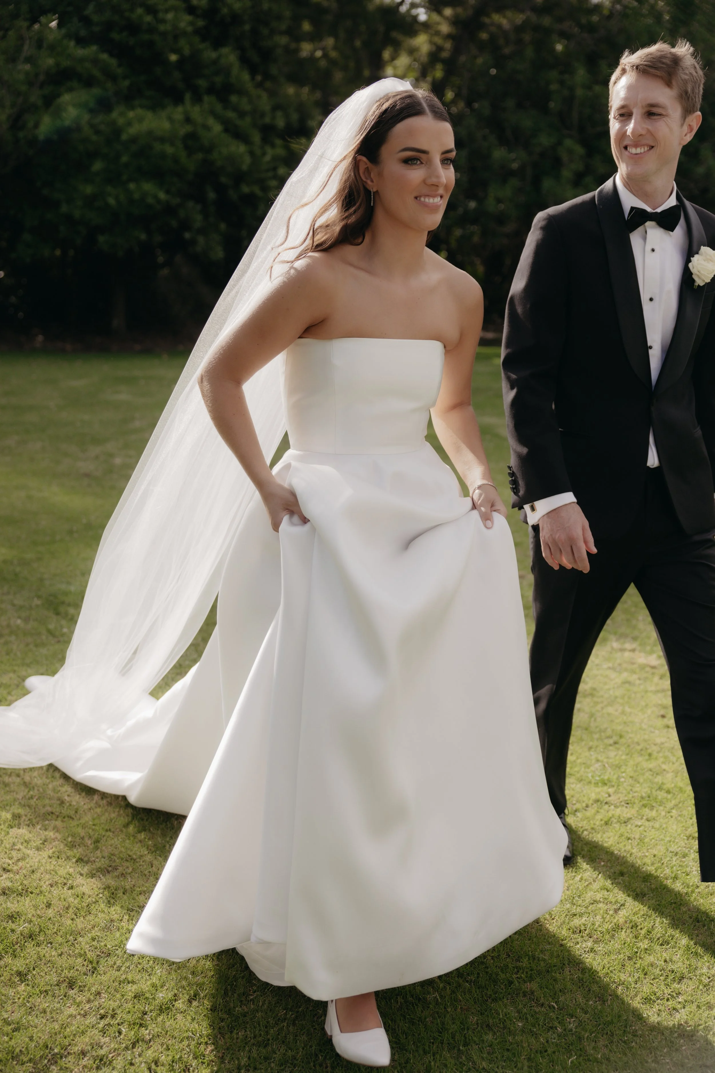 Bride in a white strapless wedding gown holding up her dress, walking outdoors with groom in a black tuxedo.