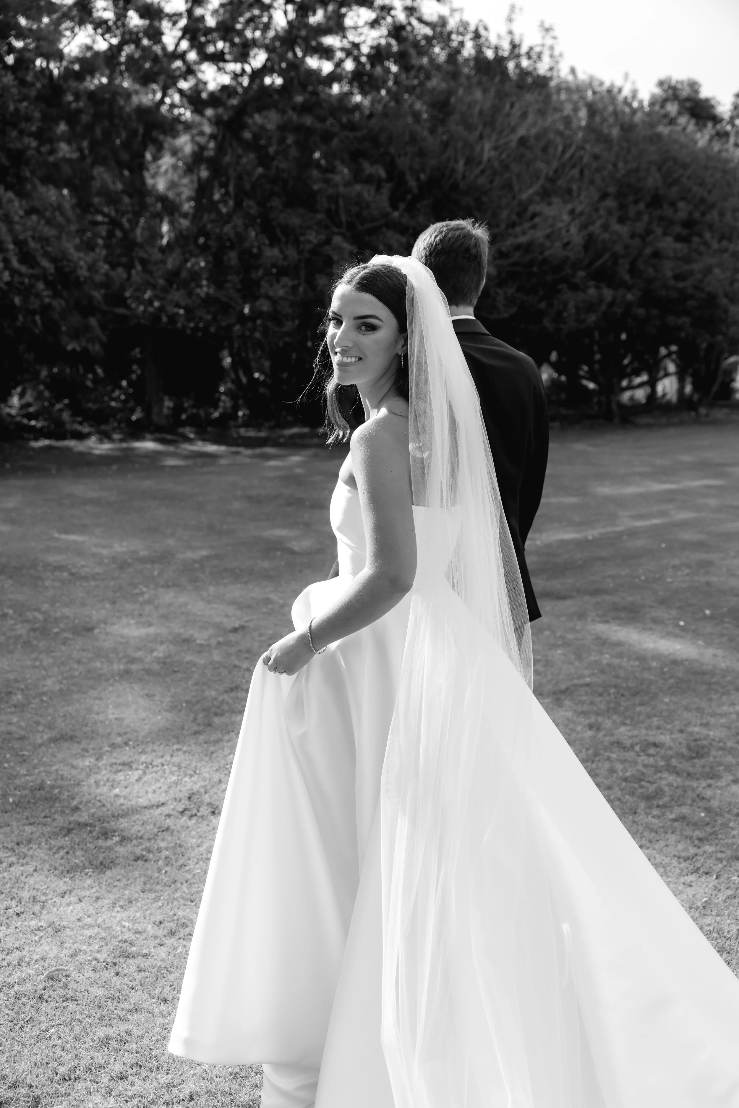 A bride in a wedding dress and veil smiling and looking back at camera, with a groom in a suit standing with his back to the camera, in an outdoor setting with trees and grass.