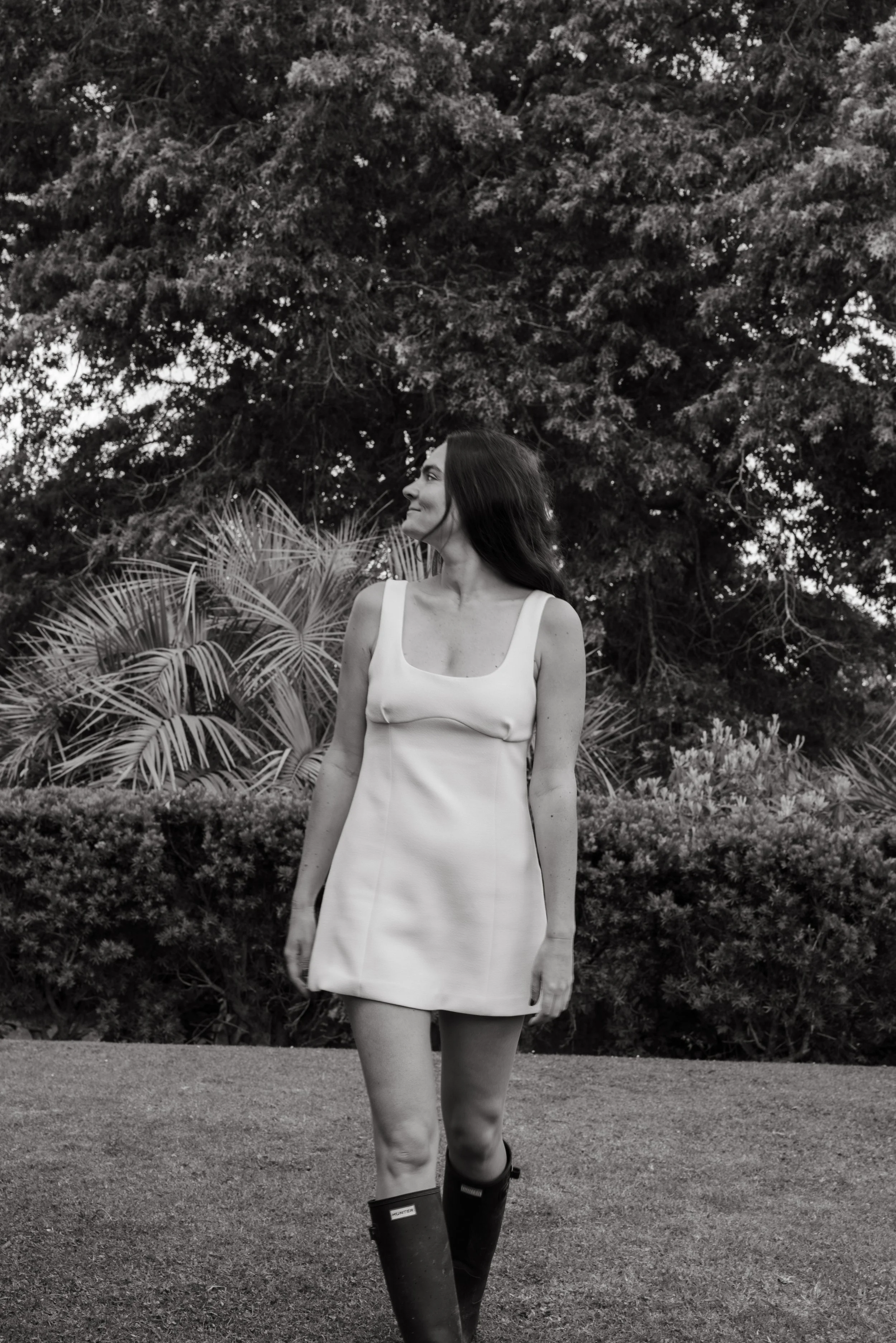 A woman in a sleeveless dress and knee-high boots standing outdoors on grass with trees and bushes in the background.