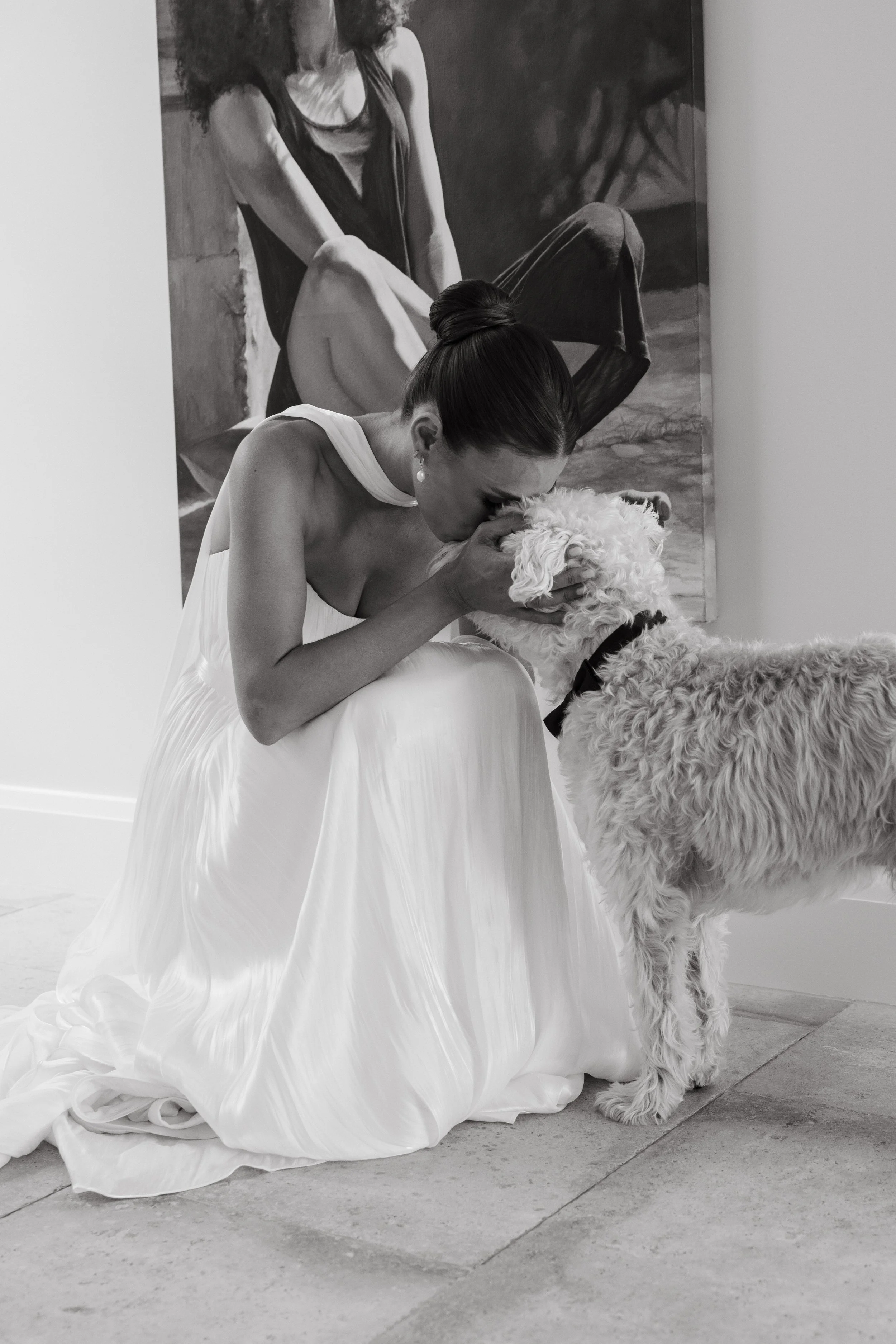 A woman in a white dress kneeling and kissing a curly-haired dog wearing a bow tie, with a portrait hanging on the wall behind her.