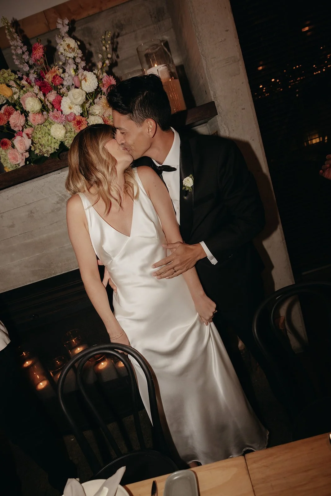 A newlywed couple sharing a kiss at their wedding celebration. The bride wears a satin white gown, and the groom is dressed in a tuxedo. They are standing in a warmly lit venue with a floral arrangement of pink and white flowers in the background.