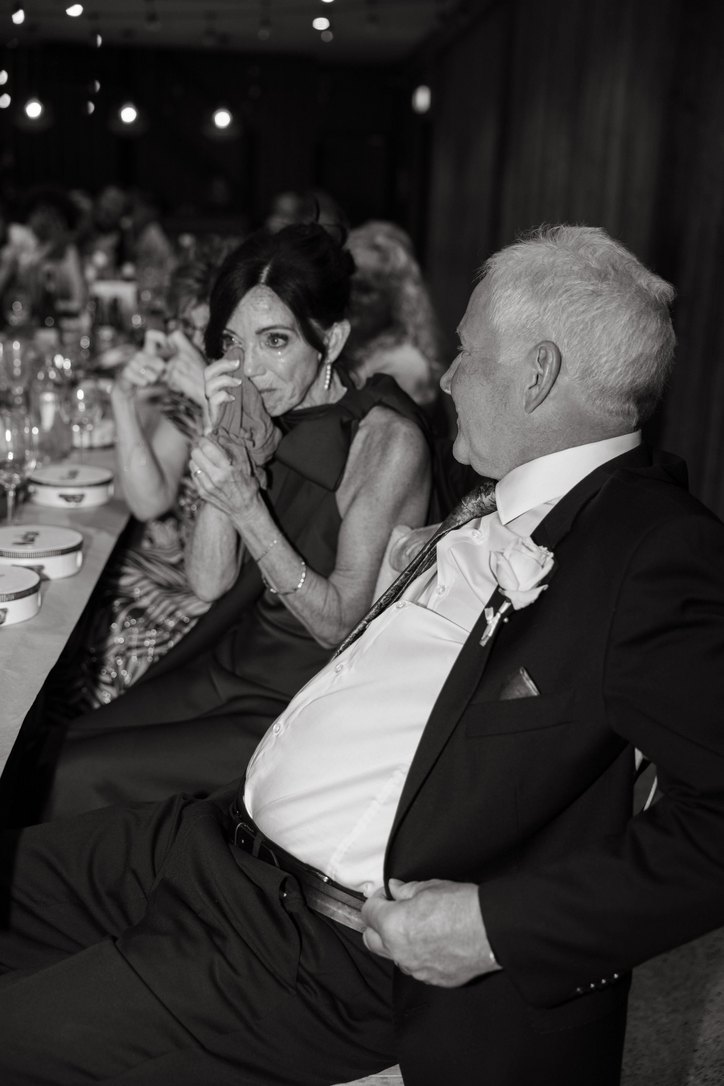 A black-and-white photo of a woman wiping tears and a man smiling at a formal event, likely a wedding reception, sitting at a long table with many guests.