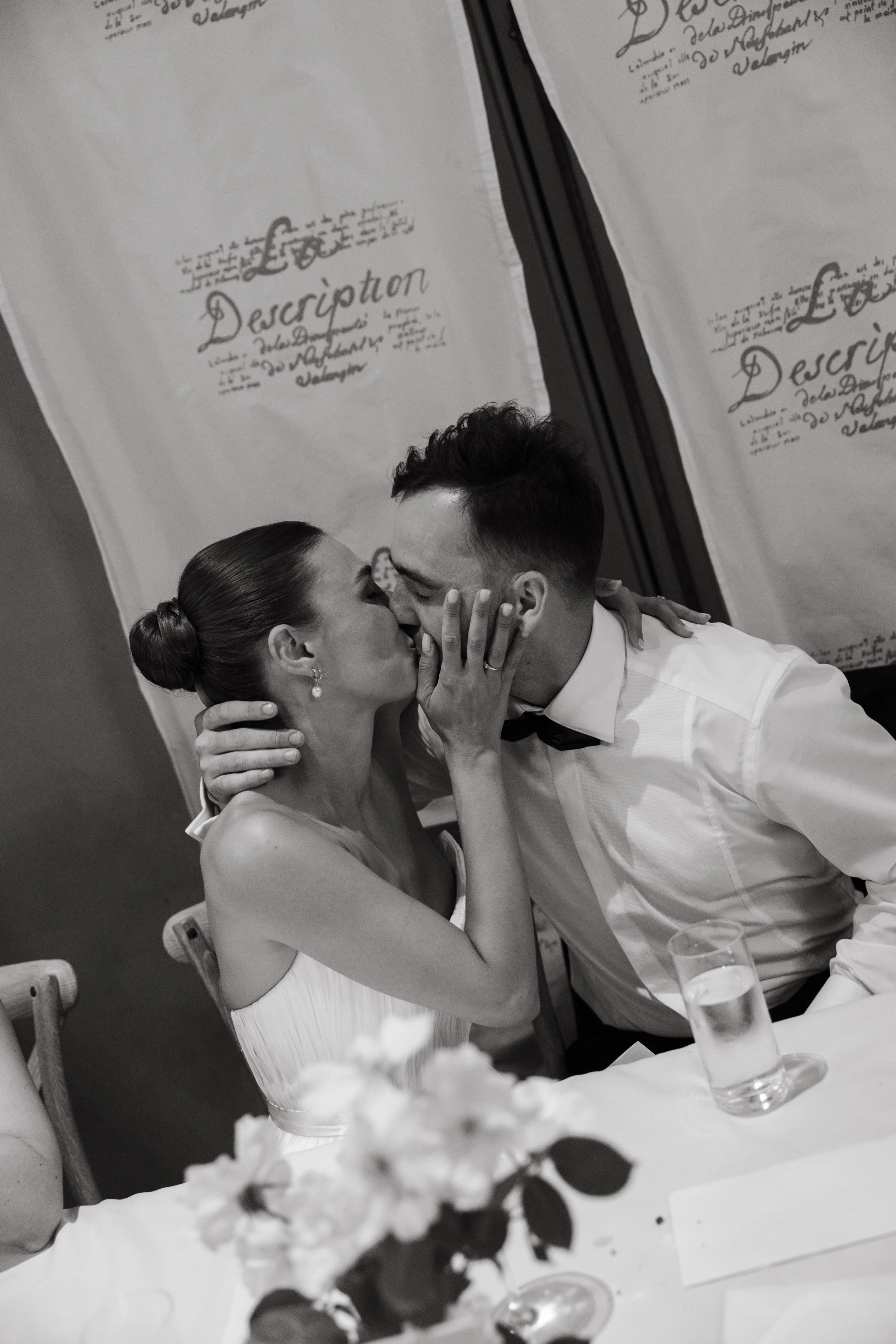 A black-and-white photo of a woman and a man kissing. The woman is wearing a strapless dress and has her hair in a bun, while the man is wearing a tuxedo with a bowtie. They are seated at a table with flowers and glasses, in a decorated setting with a backdrop that says "Love" and "Description."