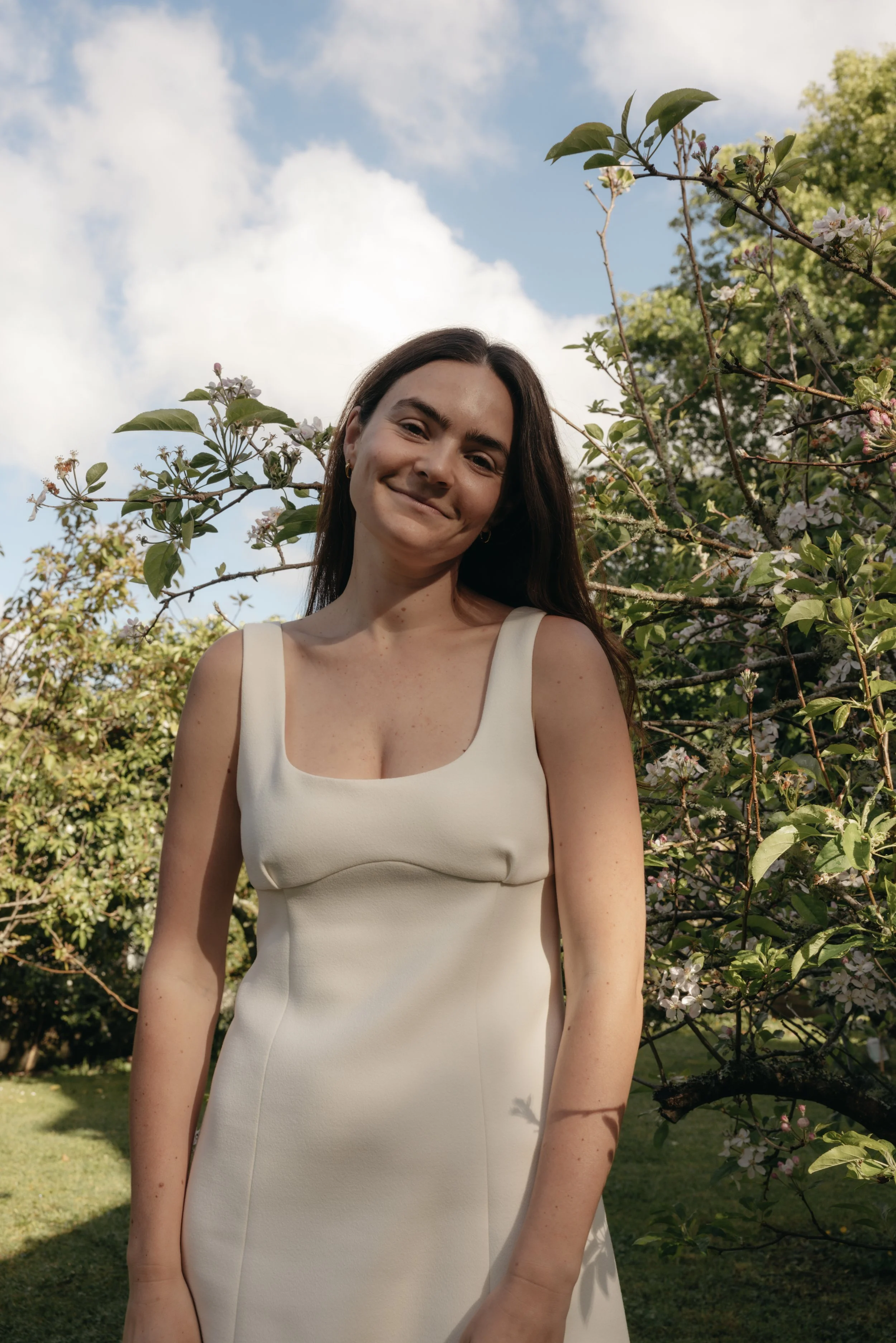 A woman with long dark hair wearing a white sleeveless dress standing outdoors in front of blooming trees.