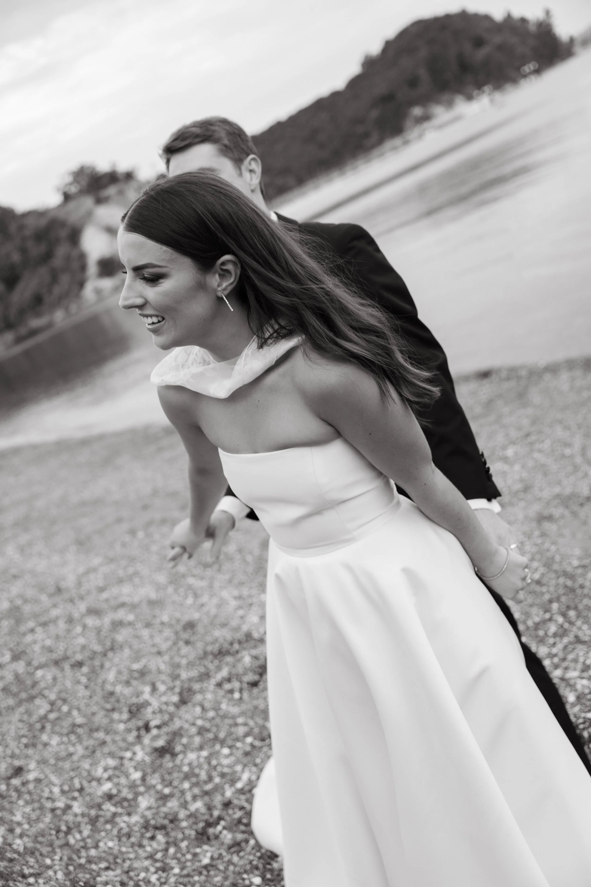 Black and white photo of a bride and groom on the beach, with the bride smiling and leaning forward, the groom standing behind her, holding her arms.