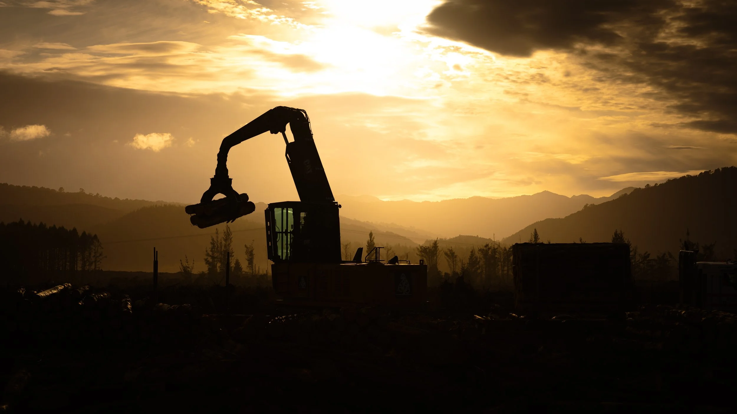 Silhouette of Caterpillar 324 construction equipment, possibly an excavator, at sunset over hilly landscape with mountains and trees in the background.