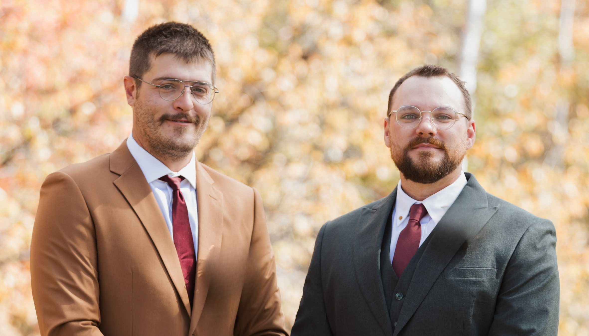 Two men dressed in suits and ties standing outdoors with autumn trees in the background.